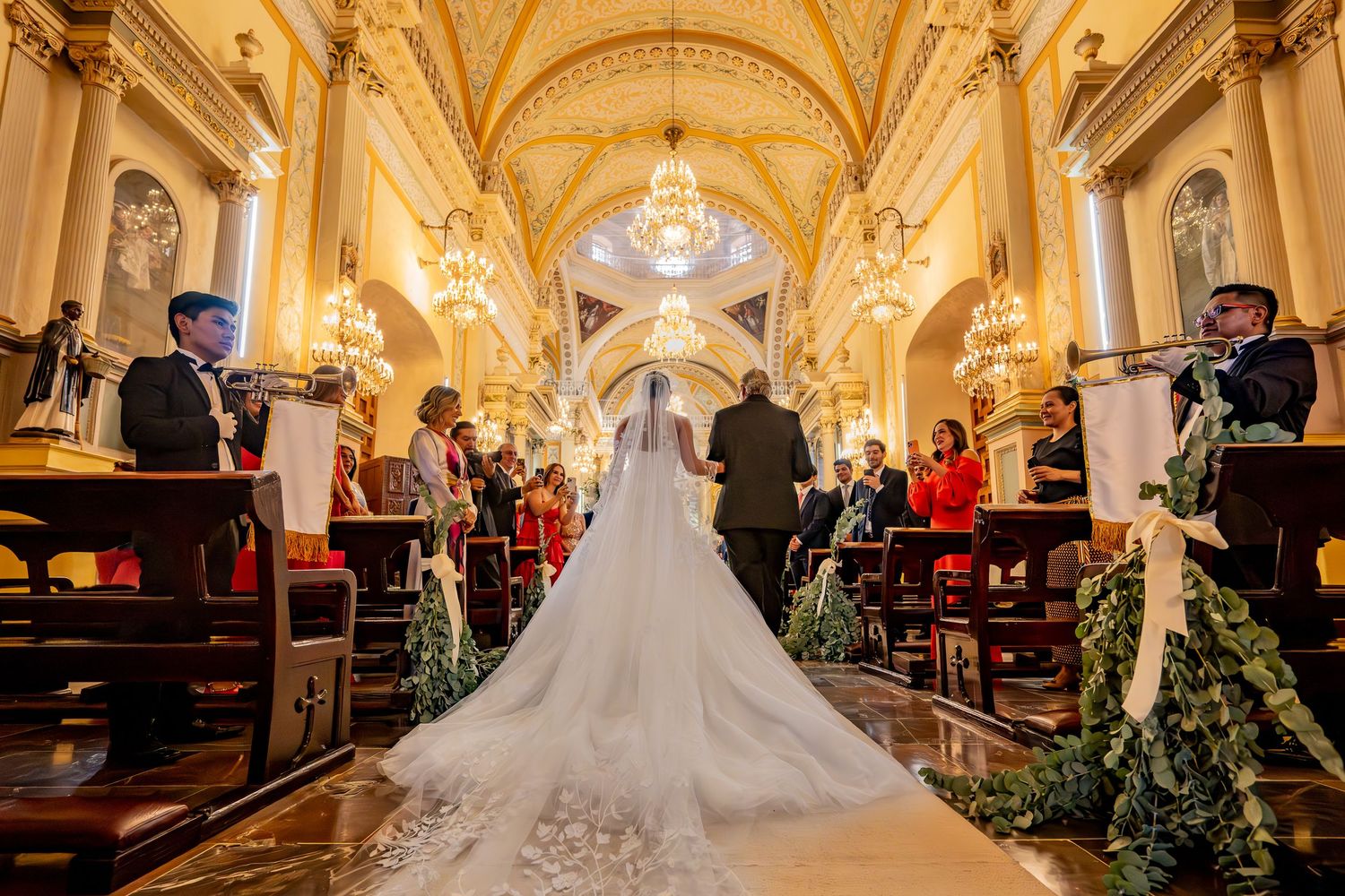 Bride in elegant white gown with long train walks down ornate church aisle during wedding ceremony.