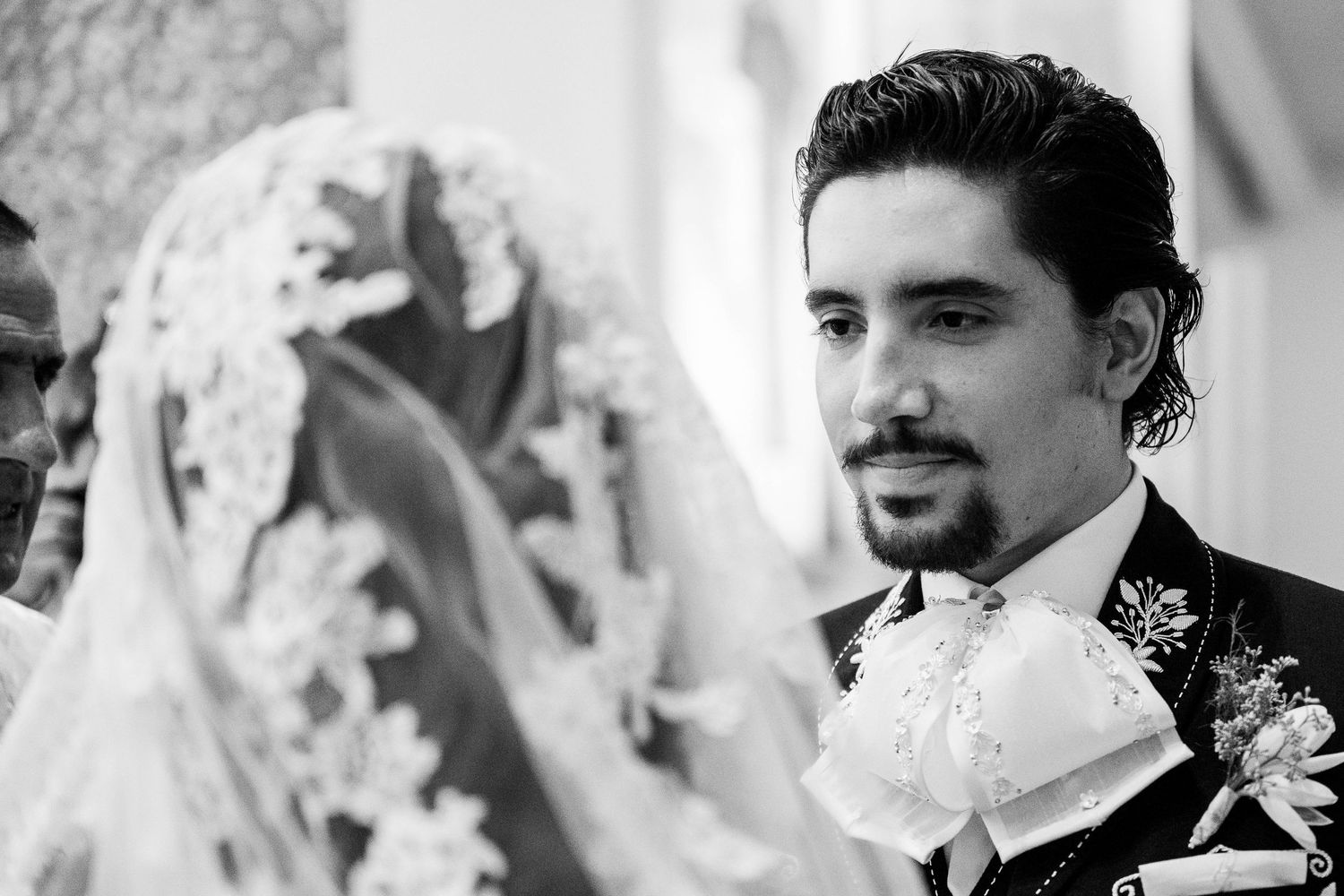 Black and white wedding photo of groom gazing at veiled bride during ceremony.