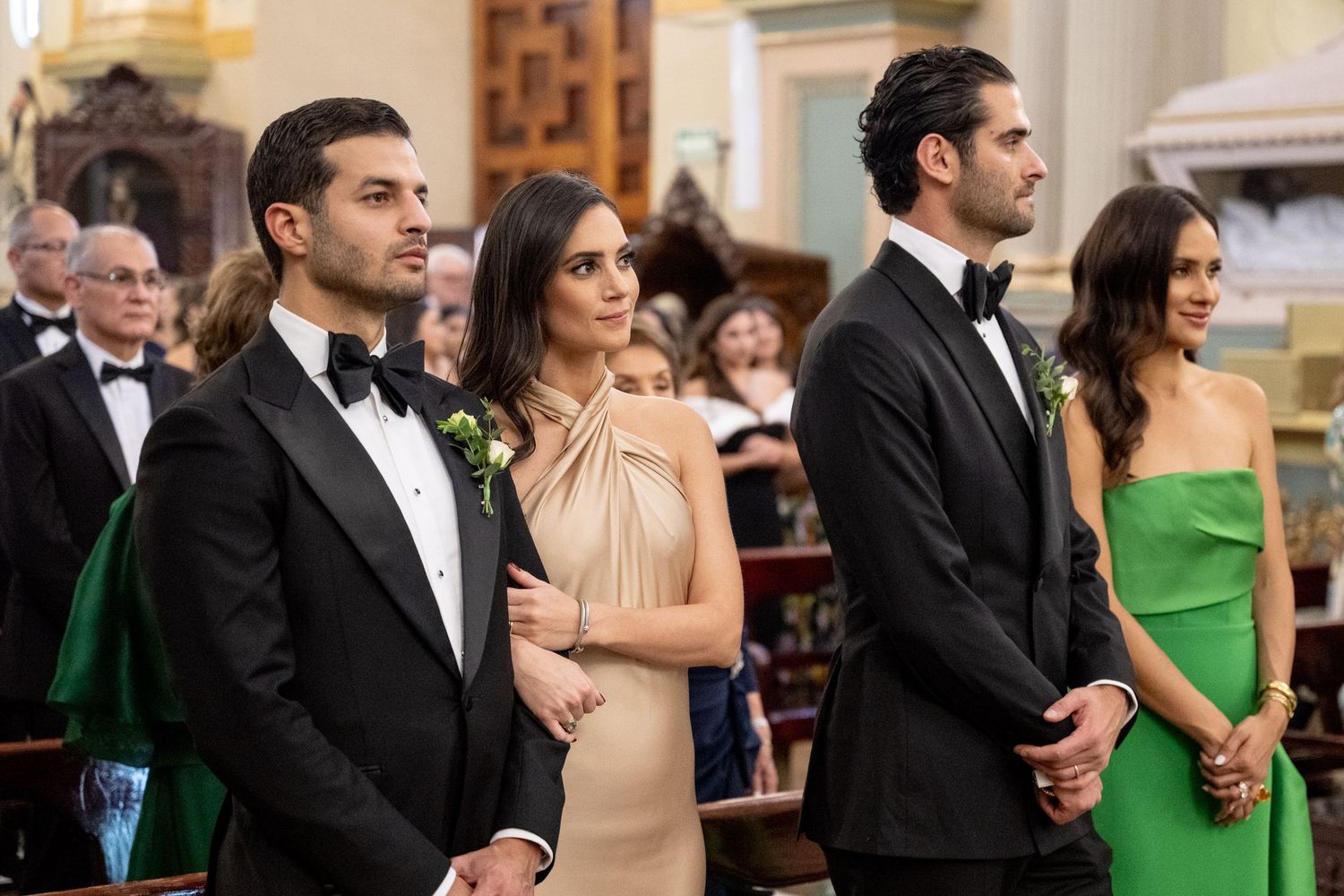 Elegantly dressed wedding guests in tuxedos and formal gowns stand in a church ceremony aisle.