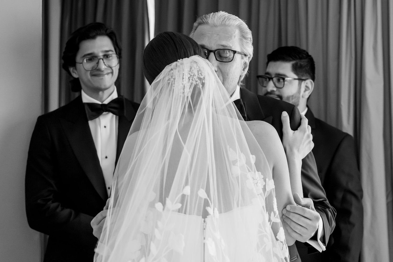 Black and white wedding photo of bride in lace veil embraced by older man while two groomsmen look on.