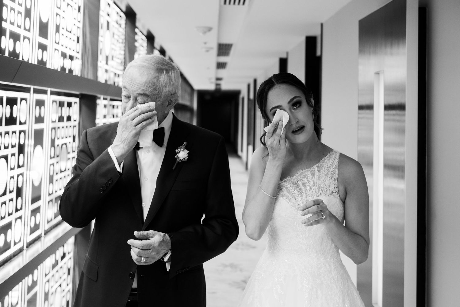 Black and white photo of a bride and older man in tuxedo both wiping tears in a hallway.