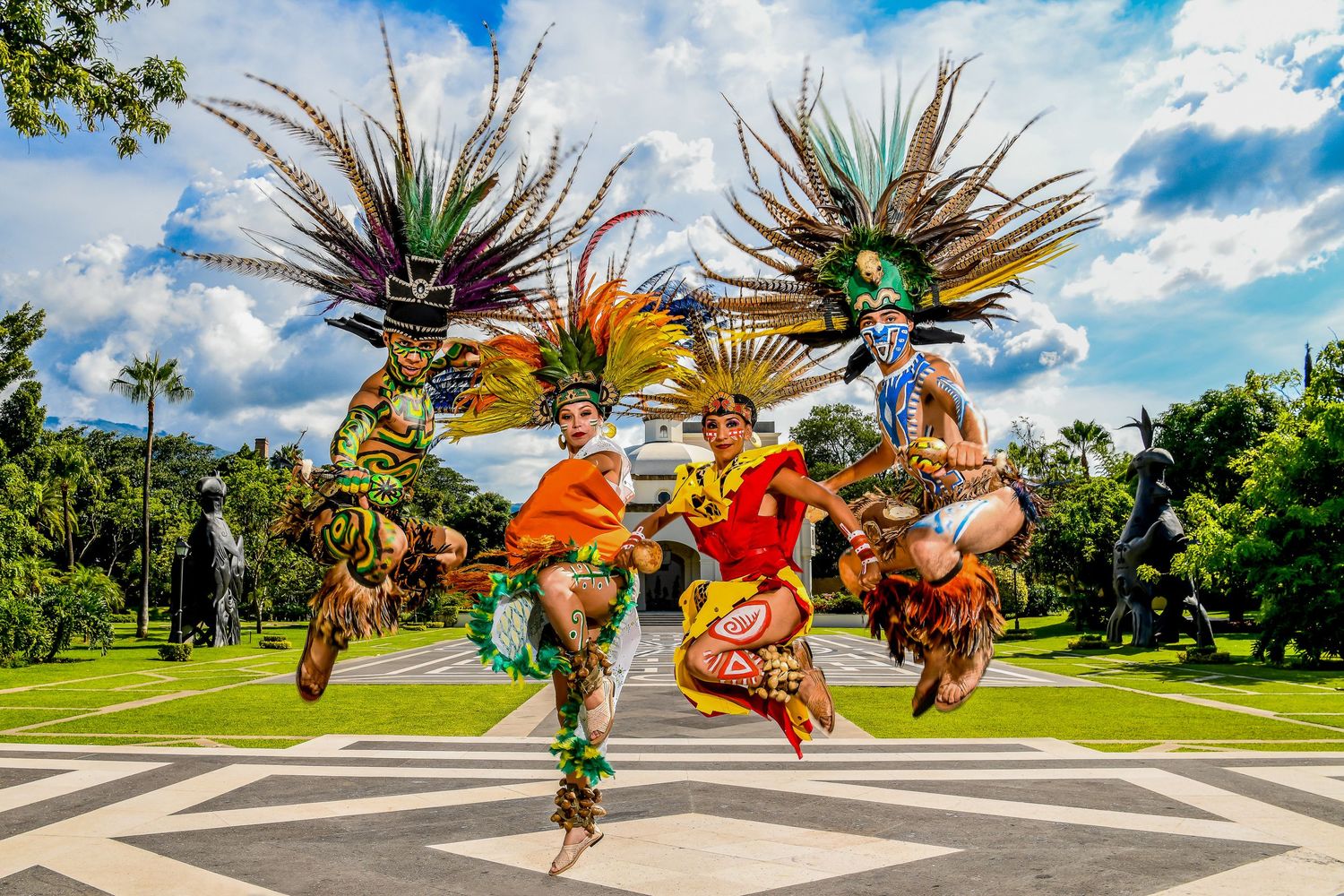 Three performers in vibrant Aztec feathered headdresses and costumes leap joyfully in an outdoor plaza.