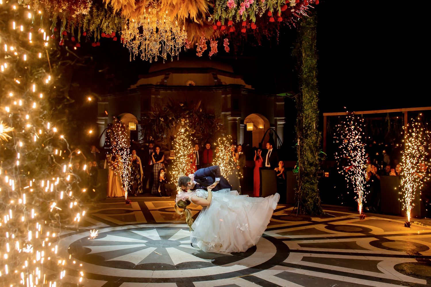Couple shares first dance under glowing fairy lights and fireworks at elegant outdoor wedding reception at night.