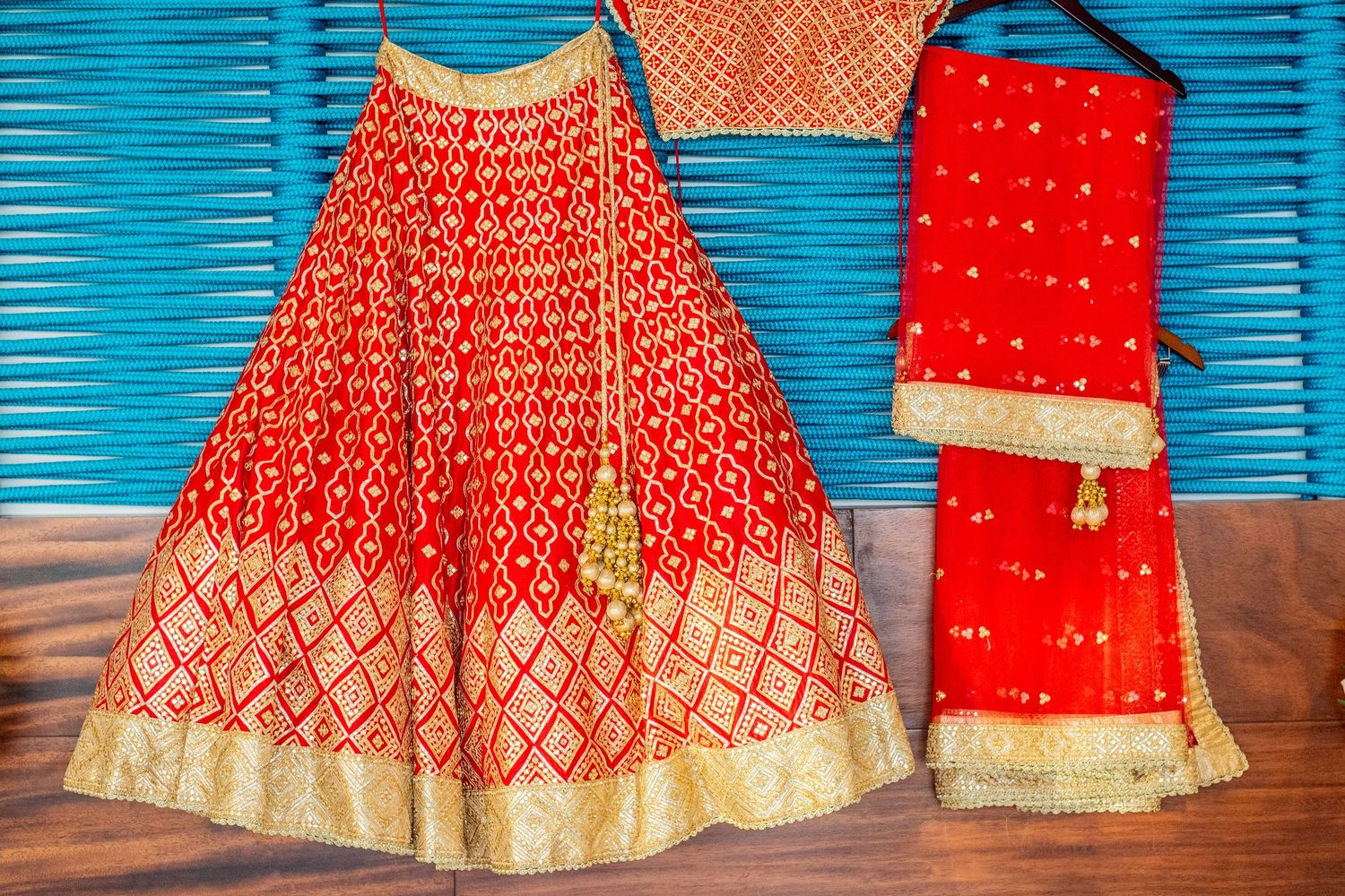 Red and gold Indian lehenga choli set with embroidered skirt, blouse, and dupatta displayed on wooden floor.