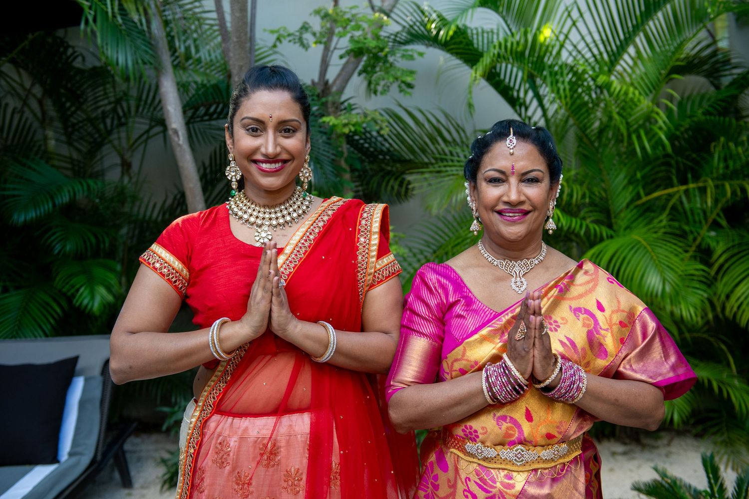 Two women in vibrant Indian sarees pose with prayer hands, smiling outdoors amid tropical greenery.