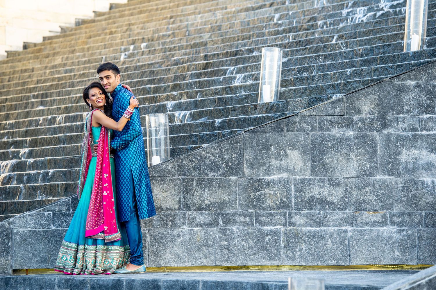Indian couple in traditional blue and pink attire embracing on stone steps at a historic monument.