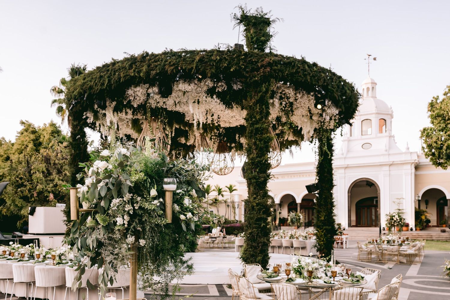 Elegant outdoor wedding reception with lush floral canopy, greenery arch, and white tablescapes near a historic venue.
