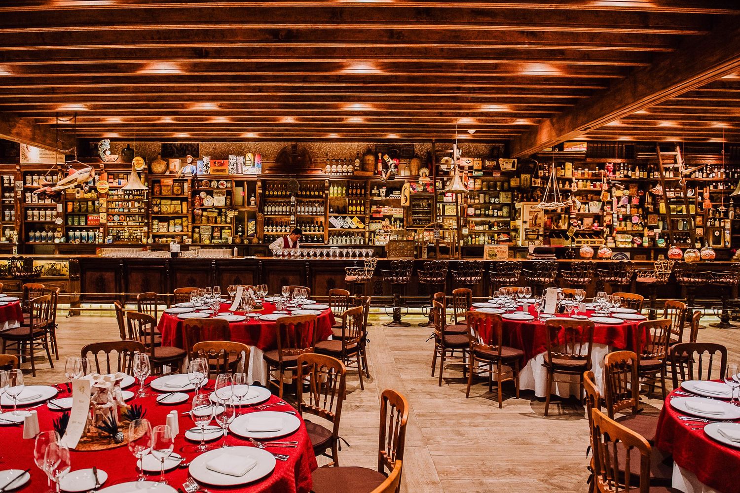Elegant restaurant dining room with red tablecloths, round tables, wooden chairs, and a well-stocked bar in the background.