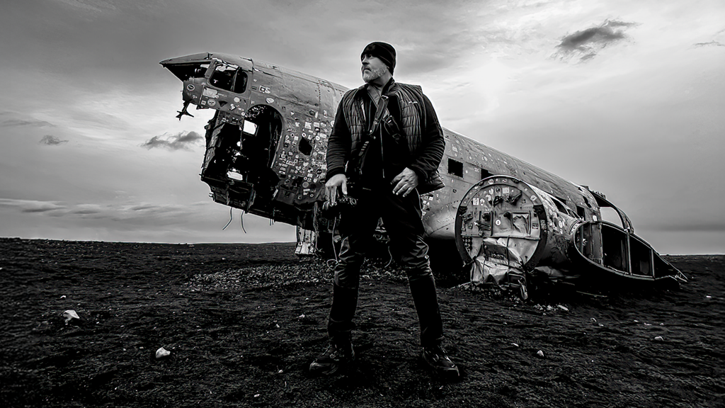 A dramatic black and white photo of a photographer standing by an abandoned airplane wreckage.