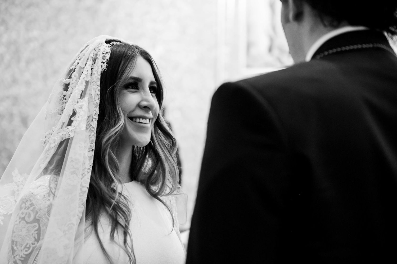 Black and white wedding photo of a smiling bride in lace veil gazing at groom during ceremony.