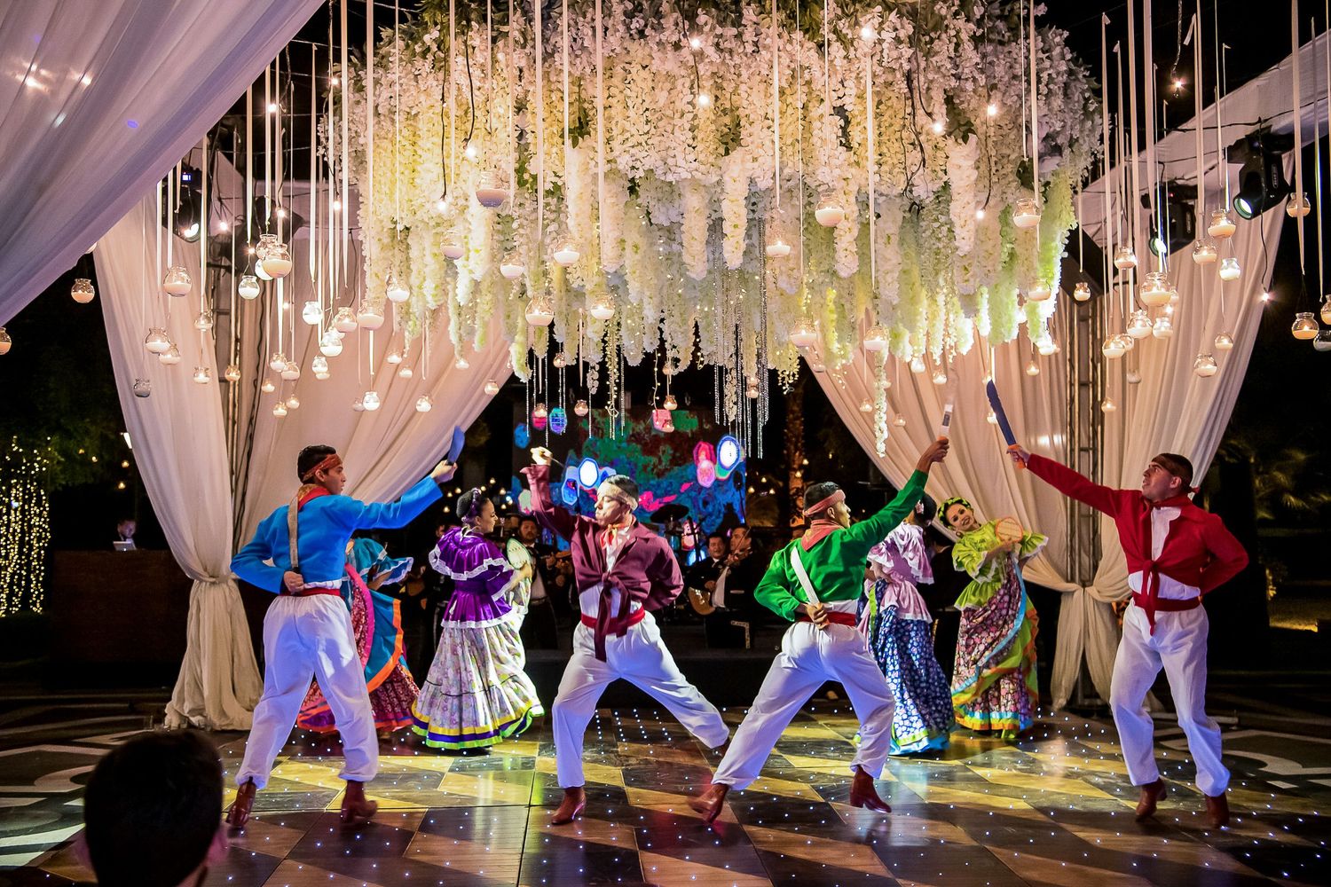Colorful dancers perform under floral chandelier at an elegant Indian wedding sangeet celebration.