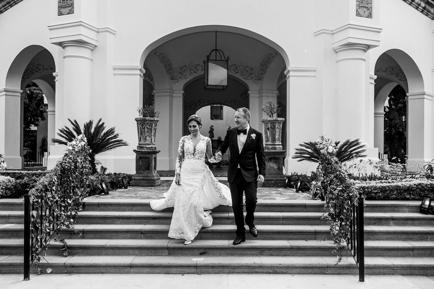 Black and white photo of bride and groom descending grand staircase of elegant wedding venue.