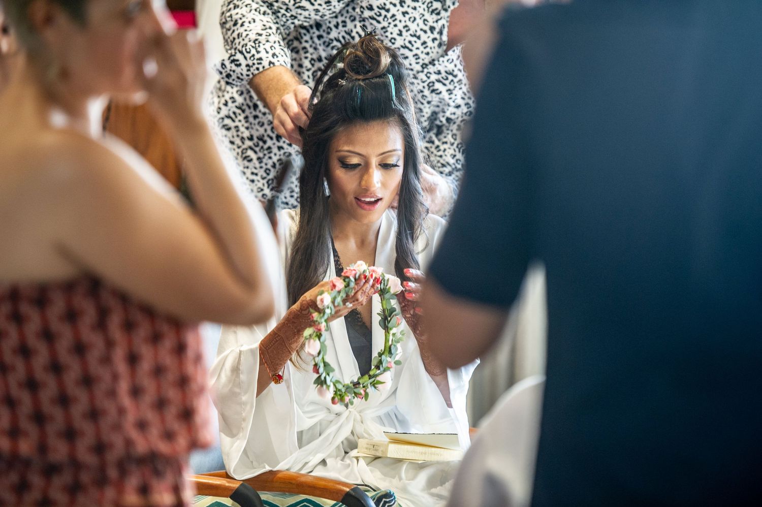 Bride in white robe holding floral crown during wedding preparation, surrounded by guests in bright natural light.