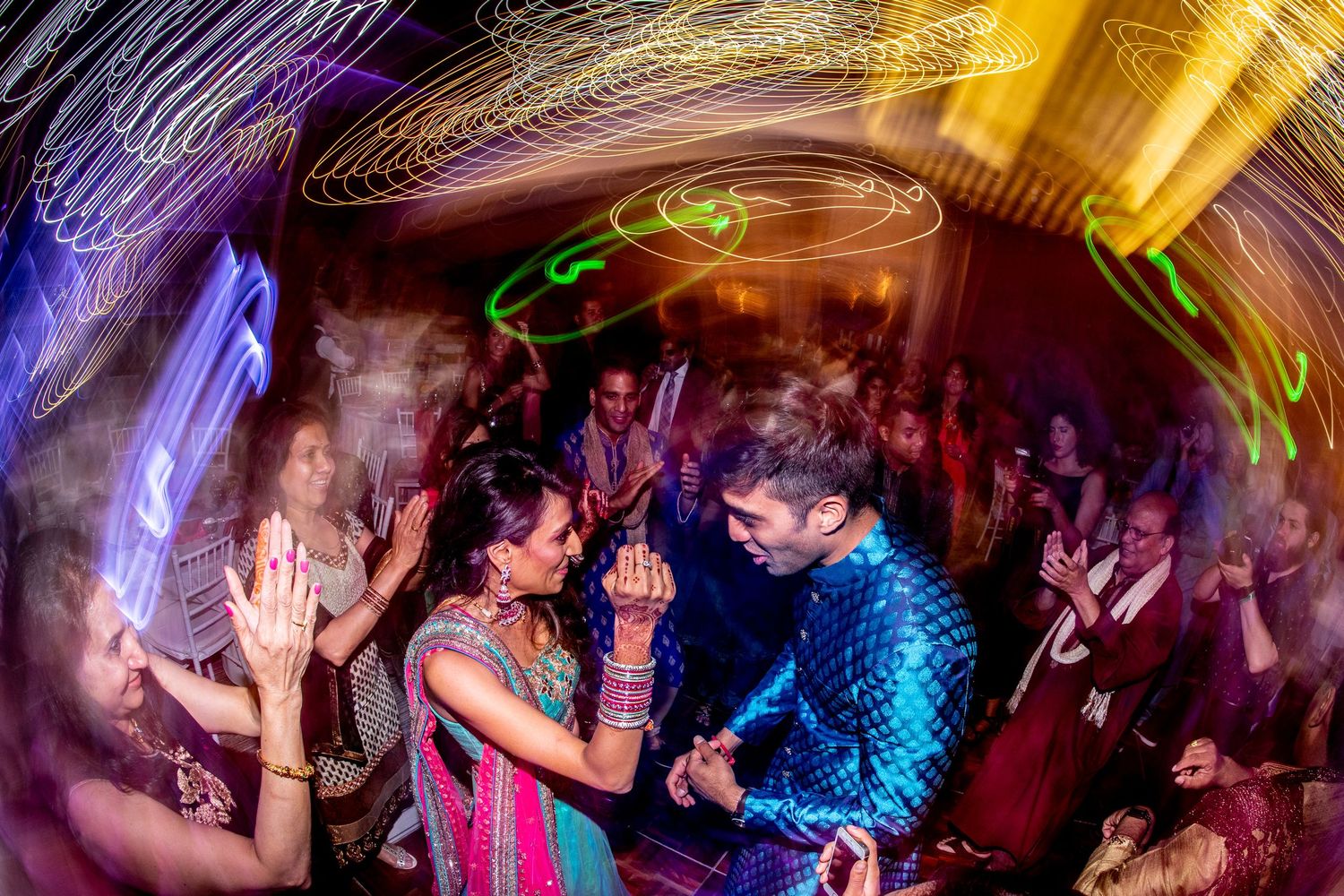 Couple dancing joyfully at a vibrant Indian wedding celebration with colorful light trails and guests clapping around them.