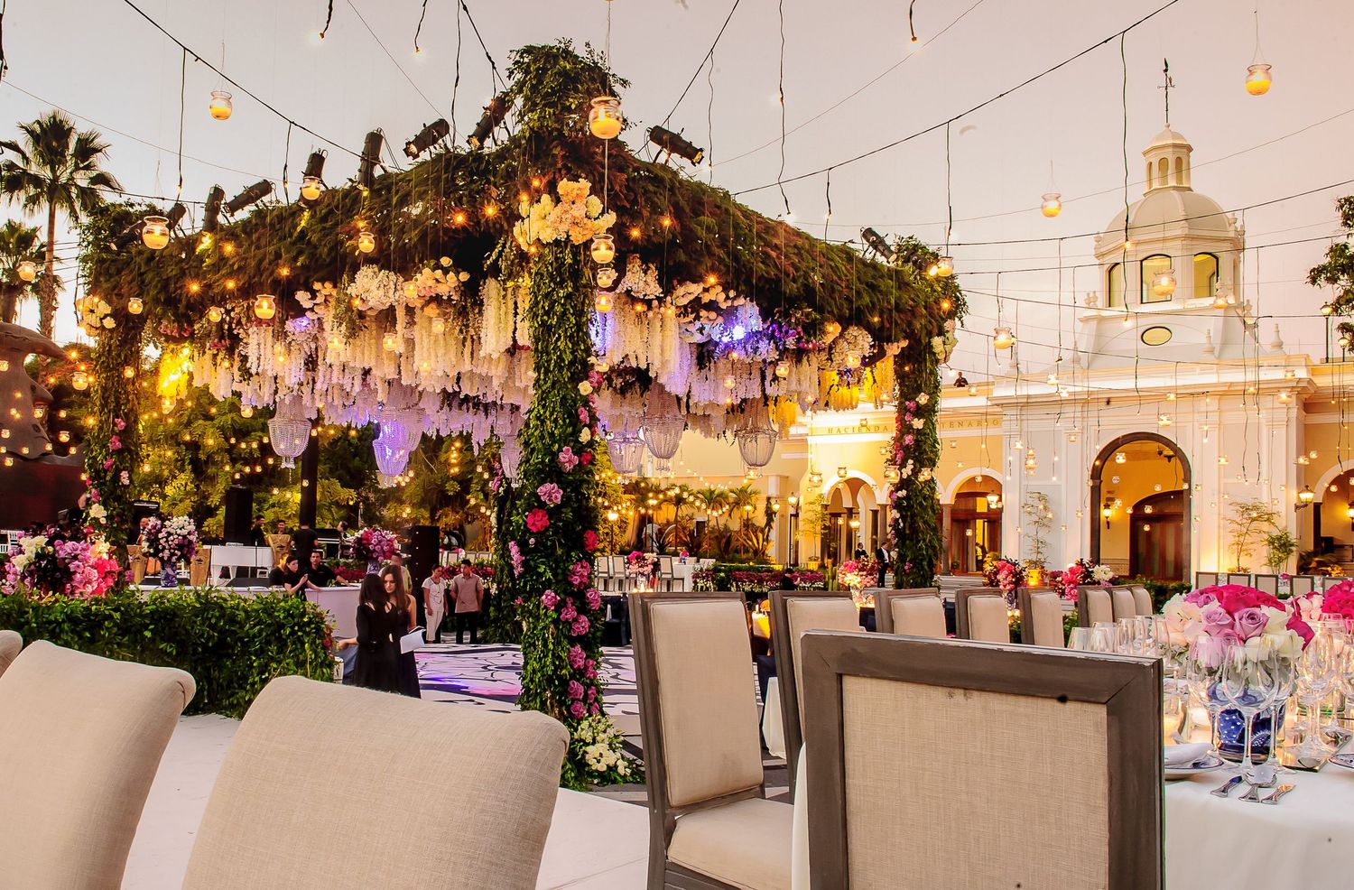 Elegant outdoor wedding reception with floral canopy, string lights, and decorated tables near a historic building.