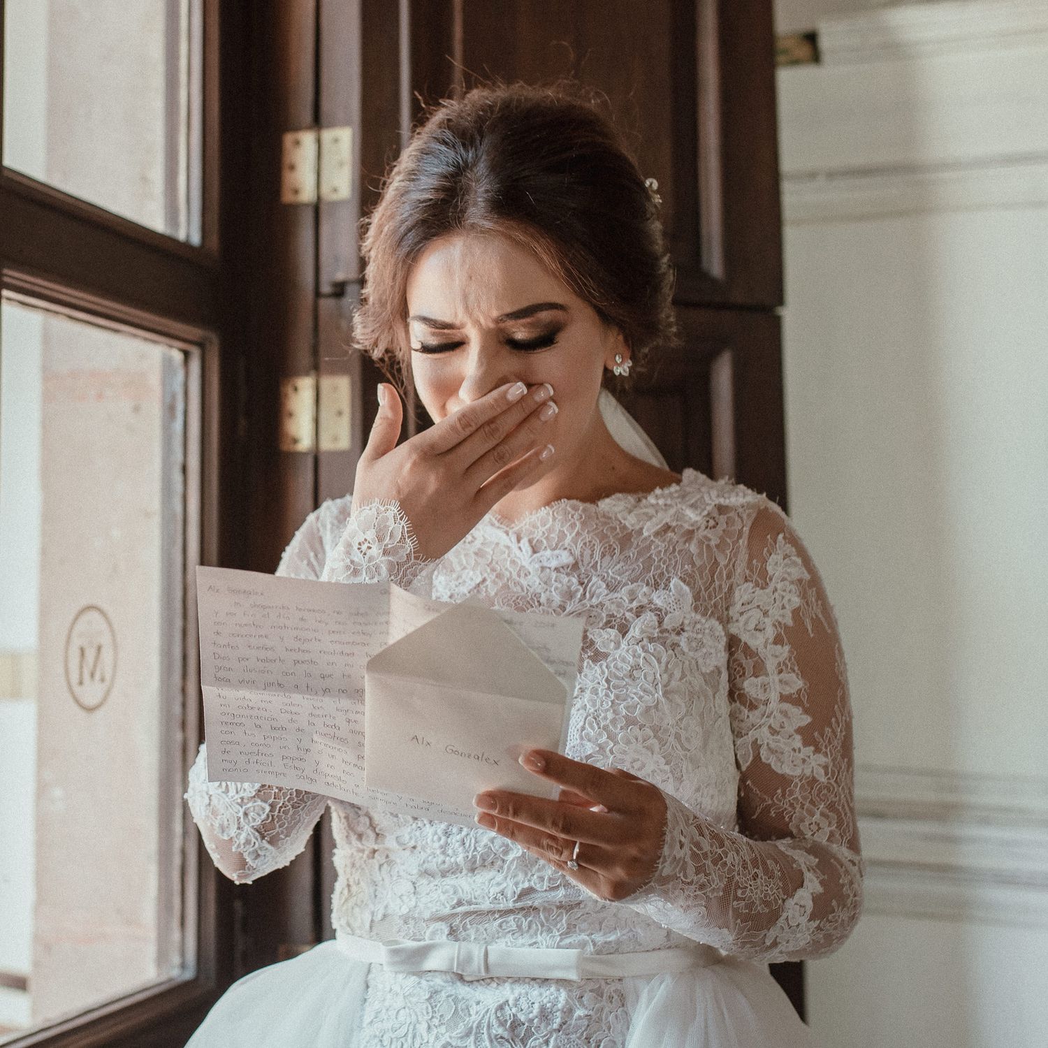 A bride in lace dress reads an emotional letter by a window.