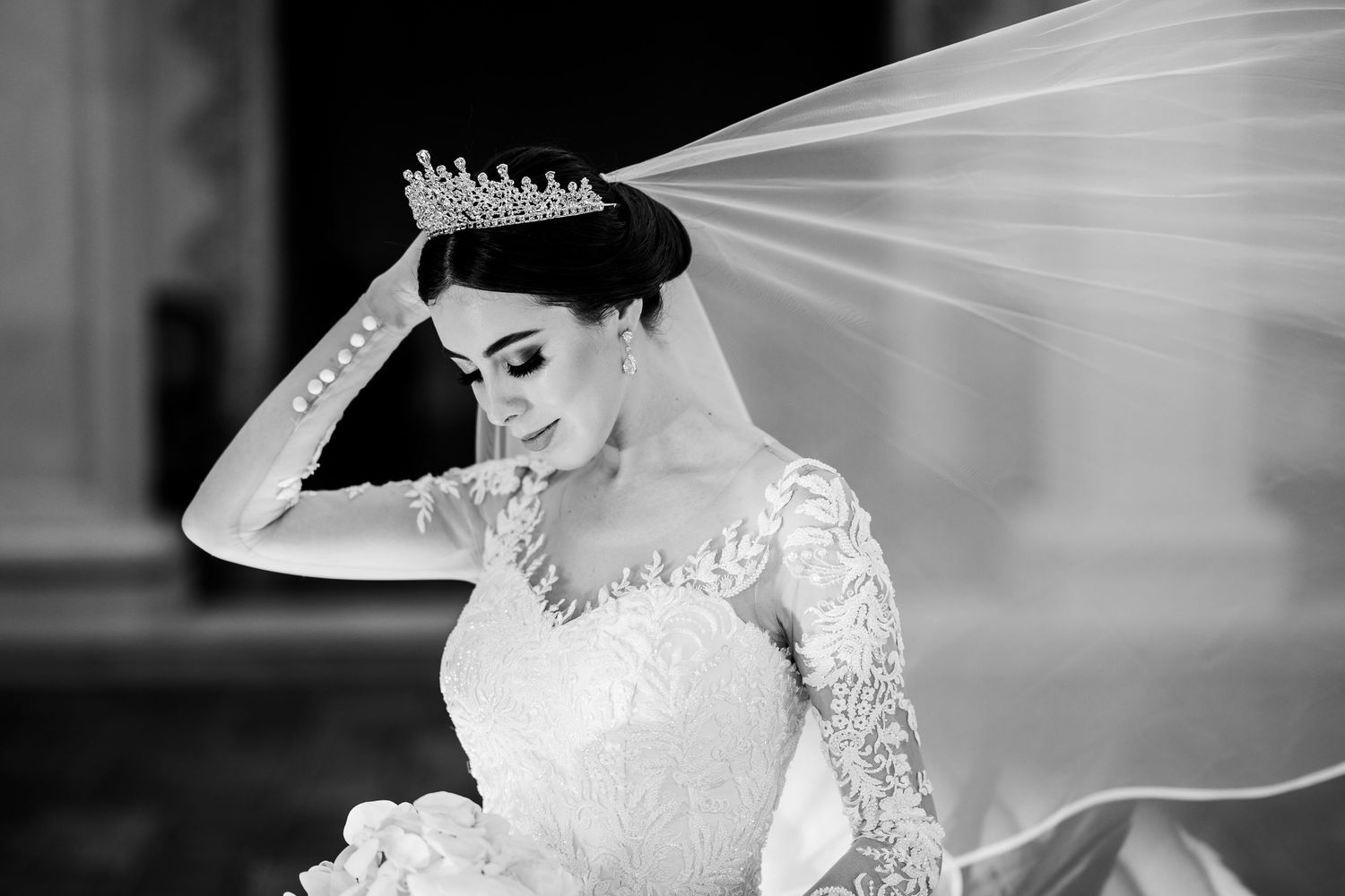 A black and white photo of a bride in long sleeve lace gown adjusting her tiara.