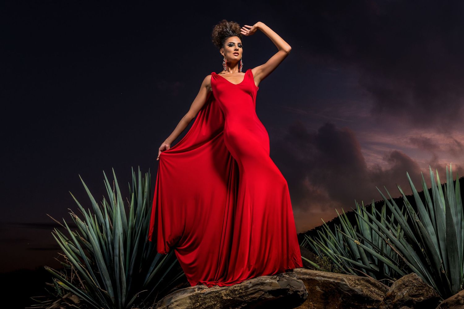 Woman in flowing red gown poses dramatically among agave plants under a stormy dark sky.
