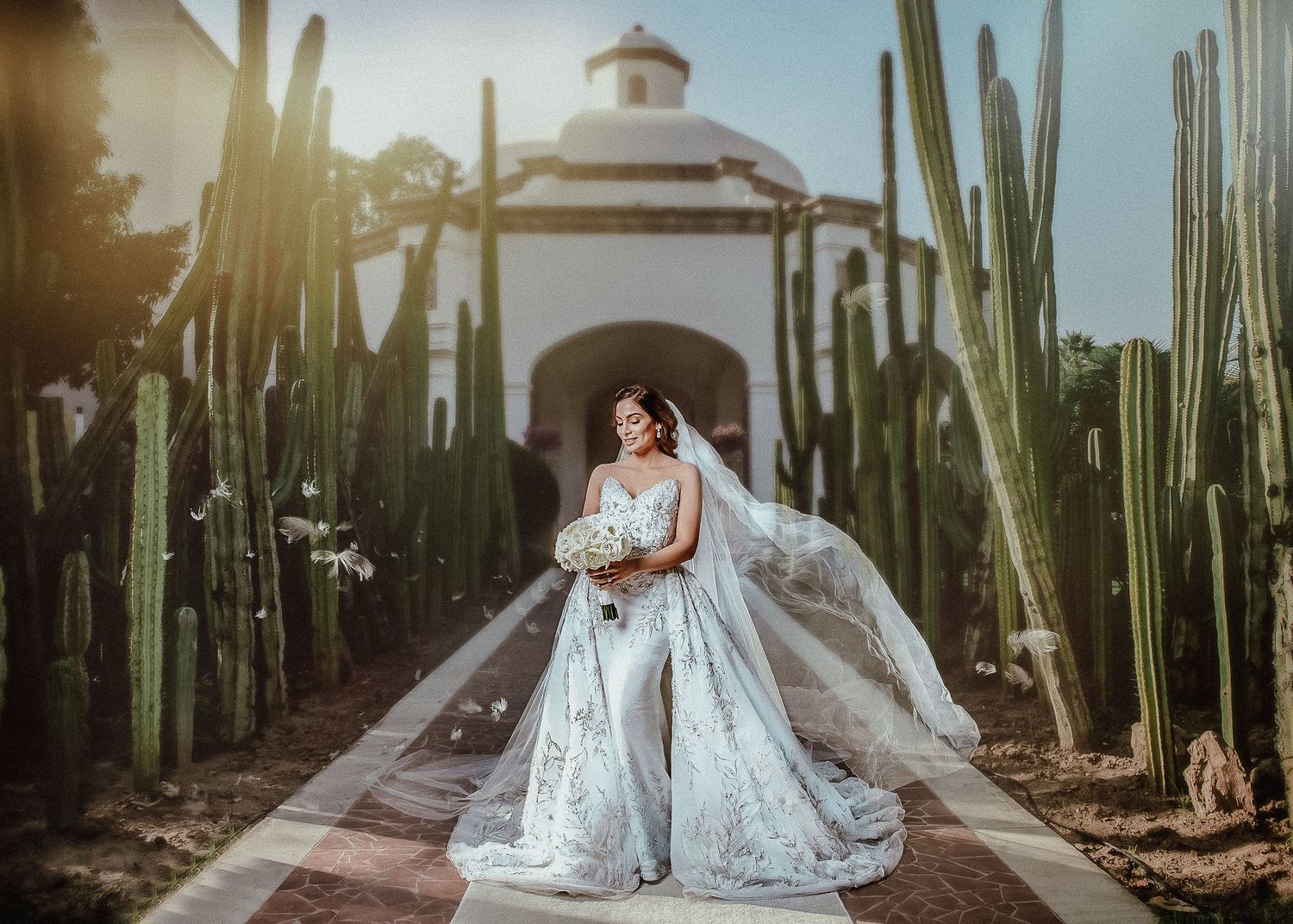 Bride in a stunning white lace gown with veil poses among tall cacti outside a white chapel.