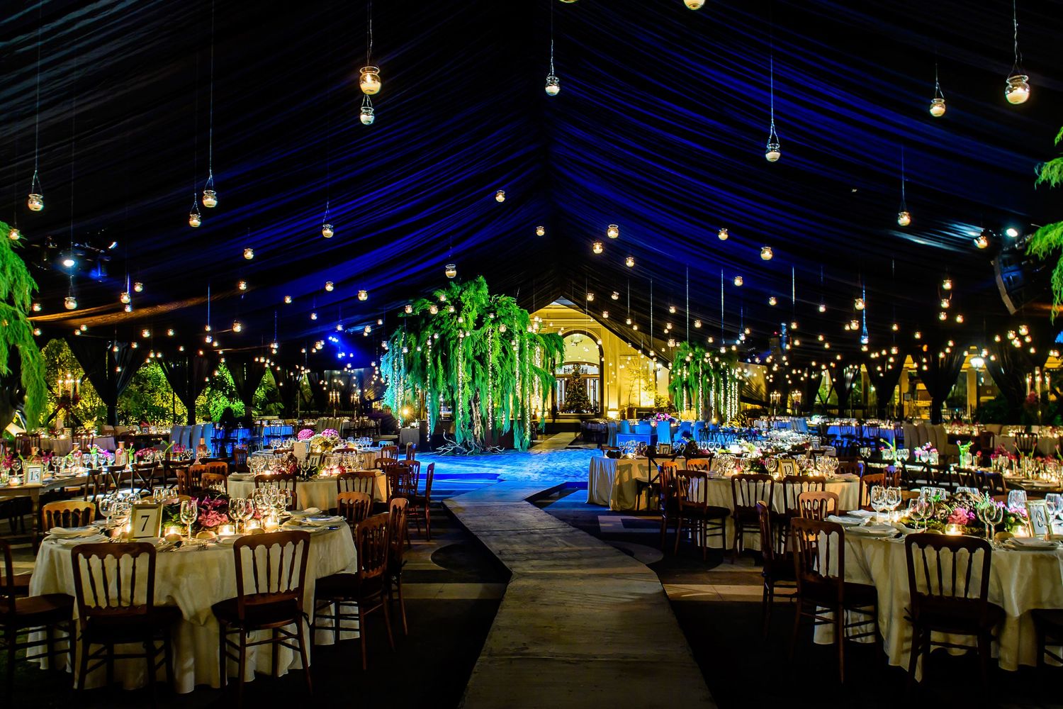 Elegant outdoor wedding reception under a tent with hanging lights, lush greenery, and beautifully decorated dining tables at night.