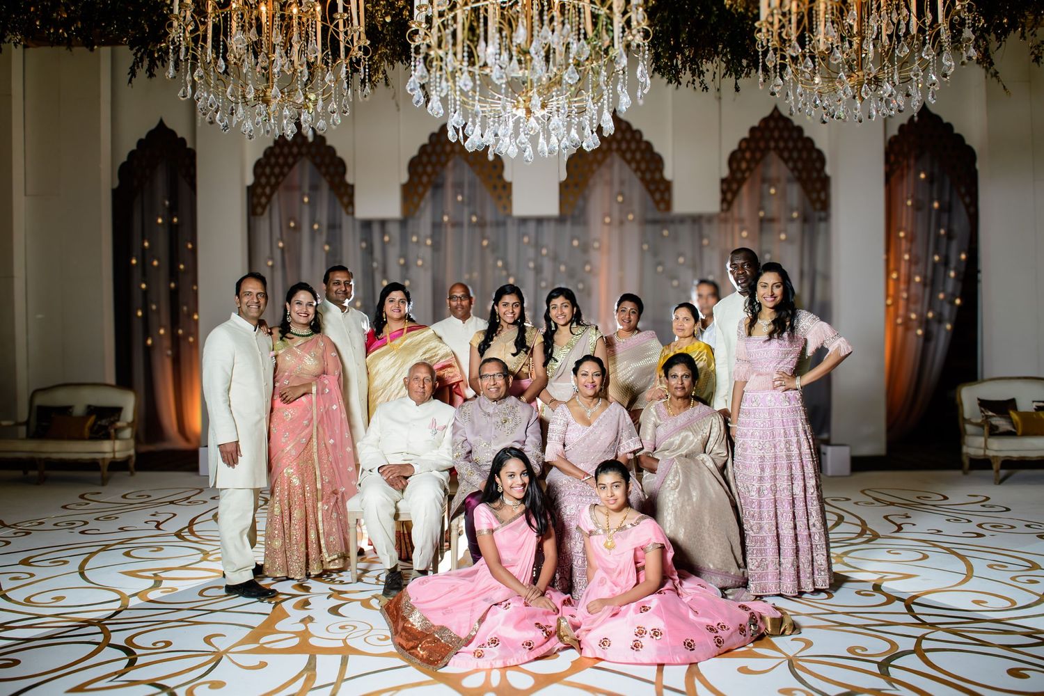 Large Indian family group photo in elegant ballroom with chandeliers, wearing traditional pink and gold festive attire.