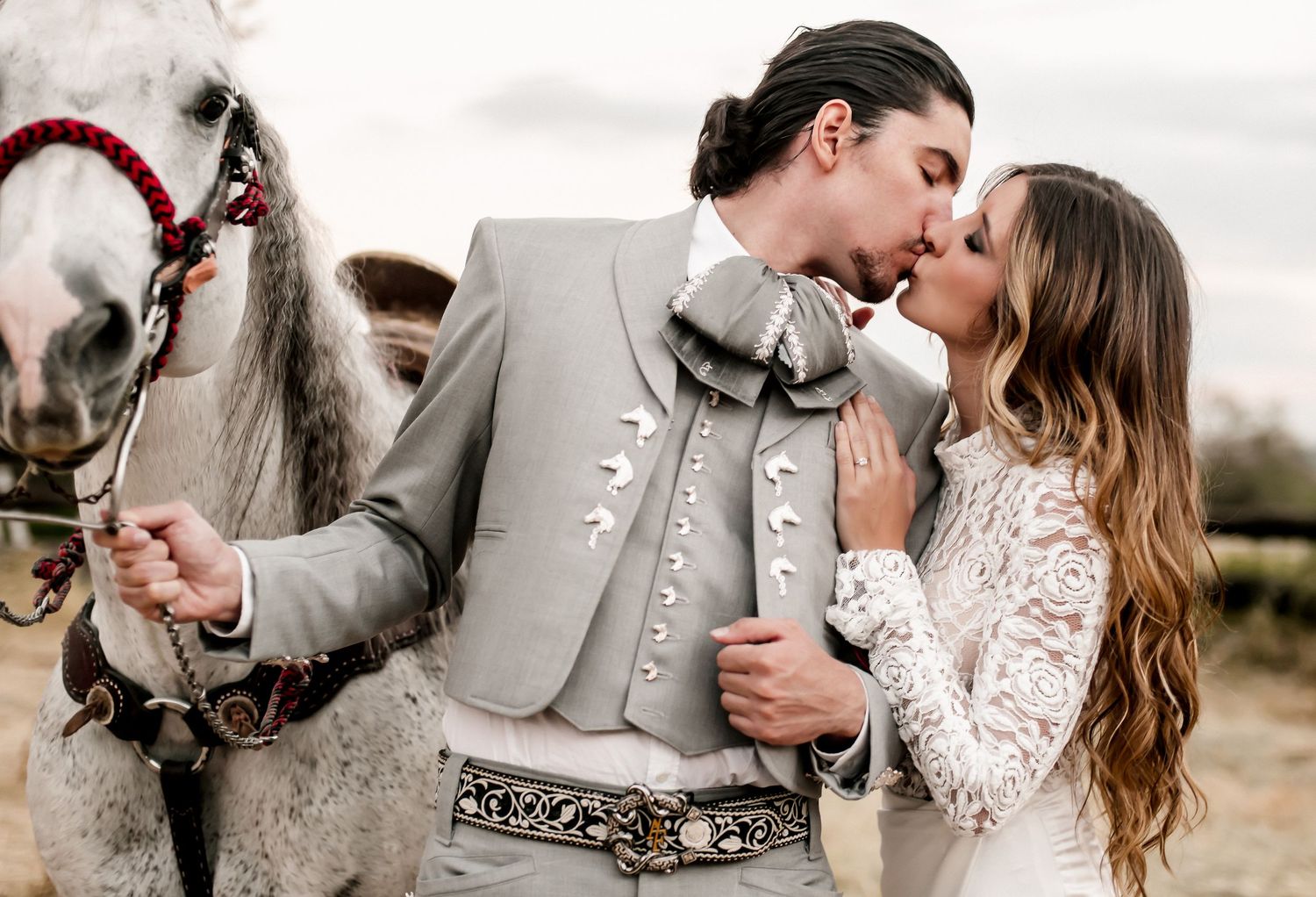 A couple in wedding attire shares a kiss beside a white horse, with the groom in a charro suit and bride in lace.