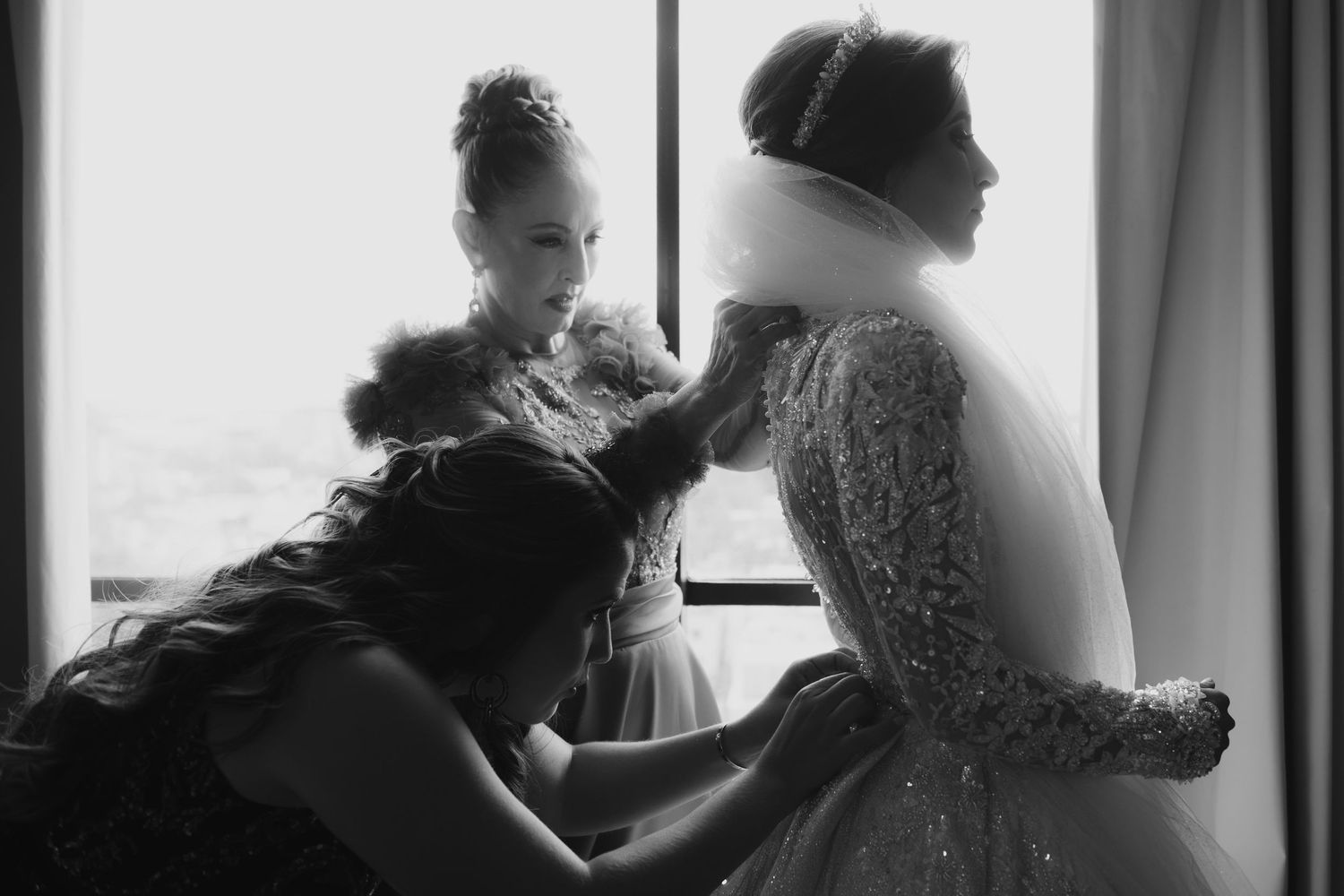 Black and white photo of a bride being helped into her wedding dress by two women near a bright window.