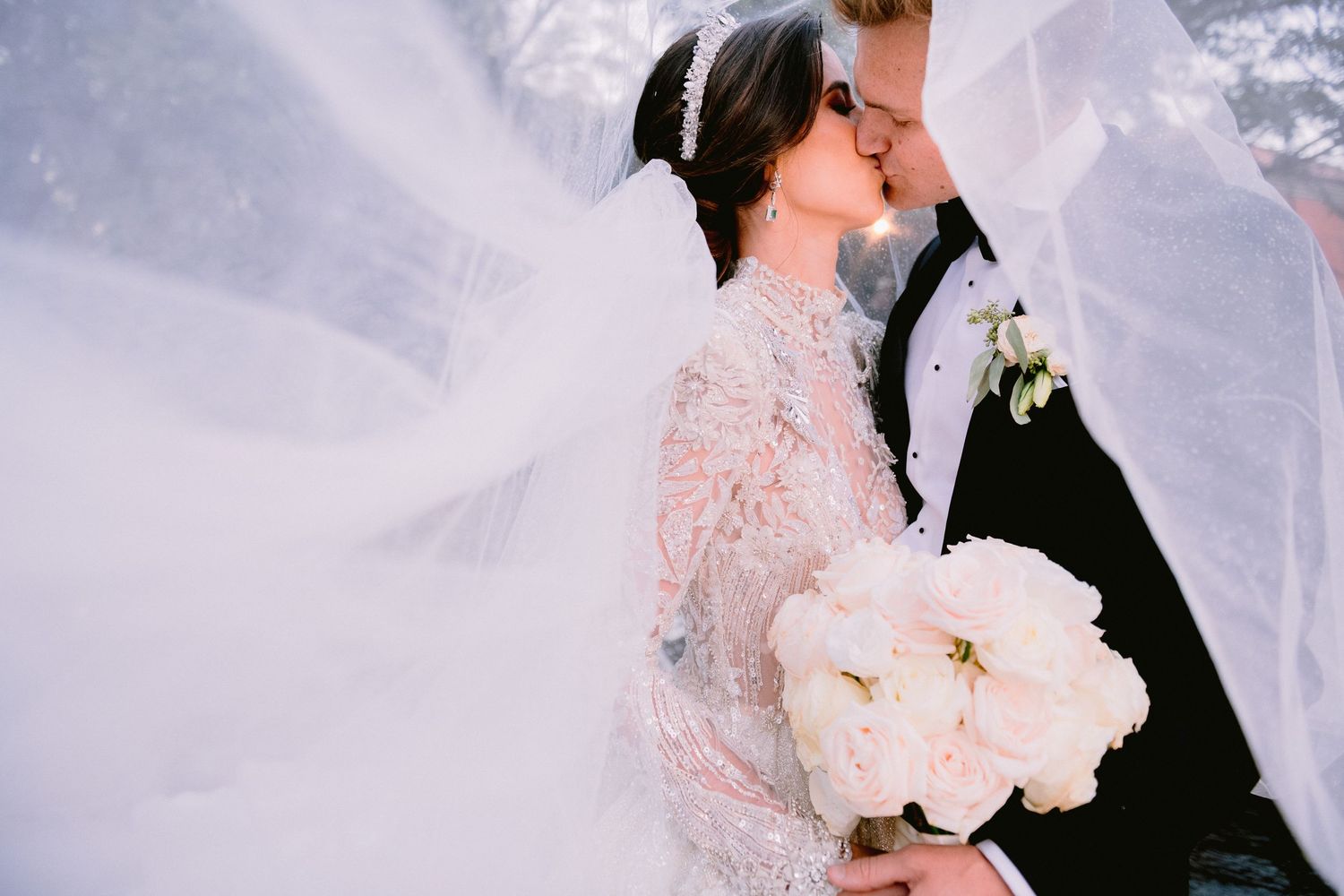 Bride and groom share a romantic kiss under flowing white veil, bride holding white peony bouquet in lace wedding gown.