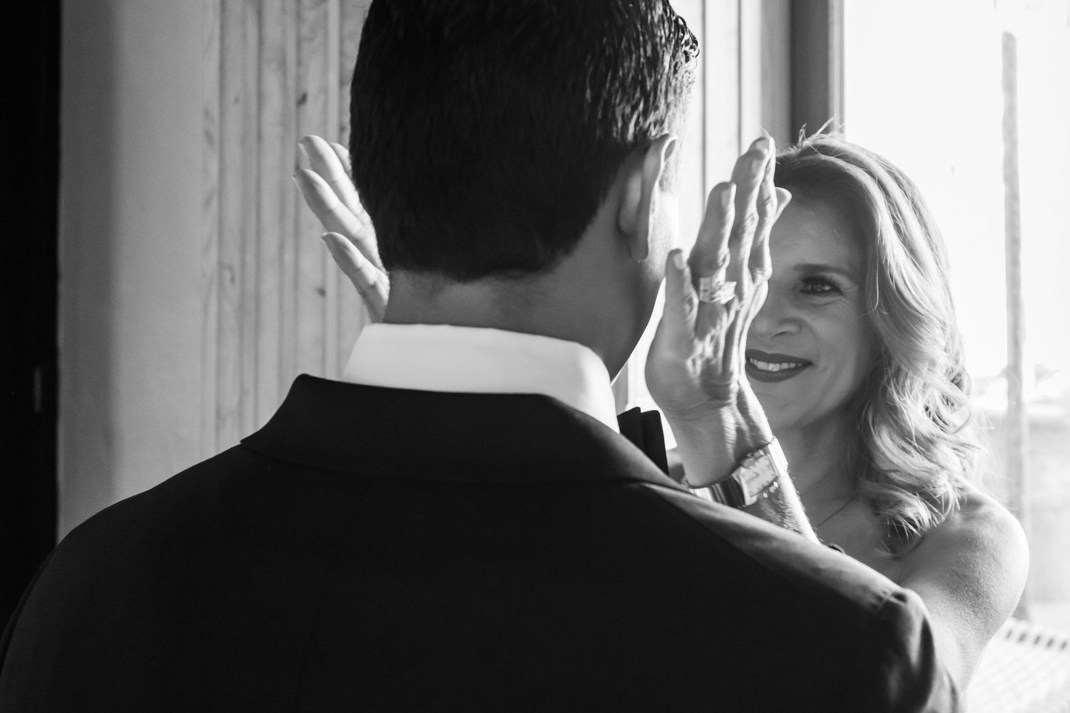 Black and white photo of a smiling blonde bride touching a groom's face near a window.