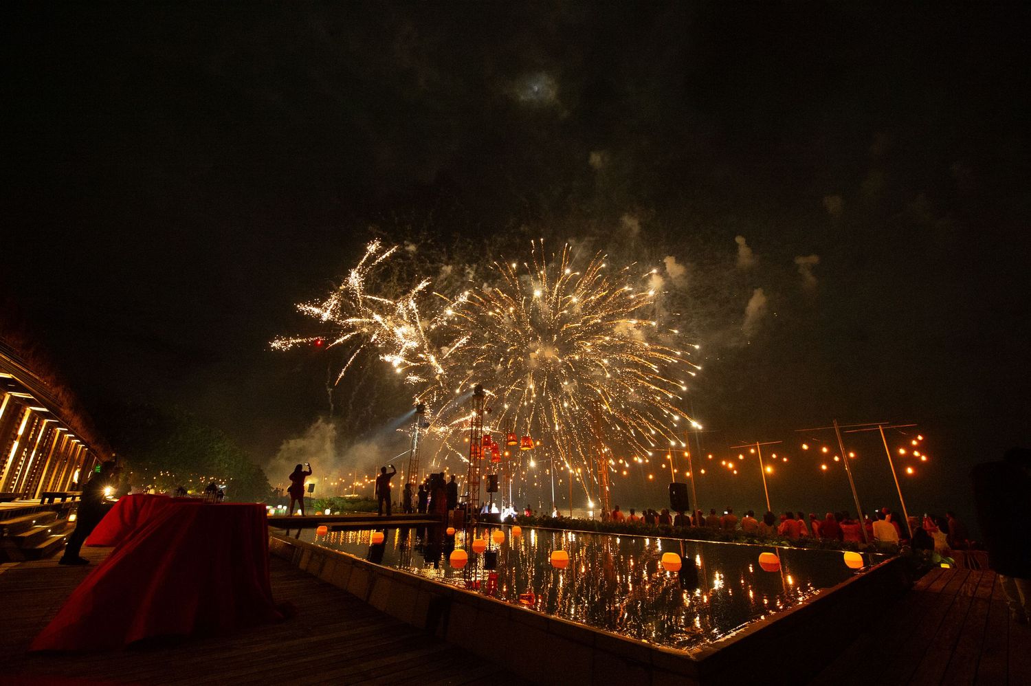 Colorful fireworks light up the night sky over a reflective pool with spectators watching the dazzling display.