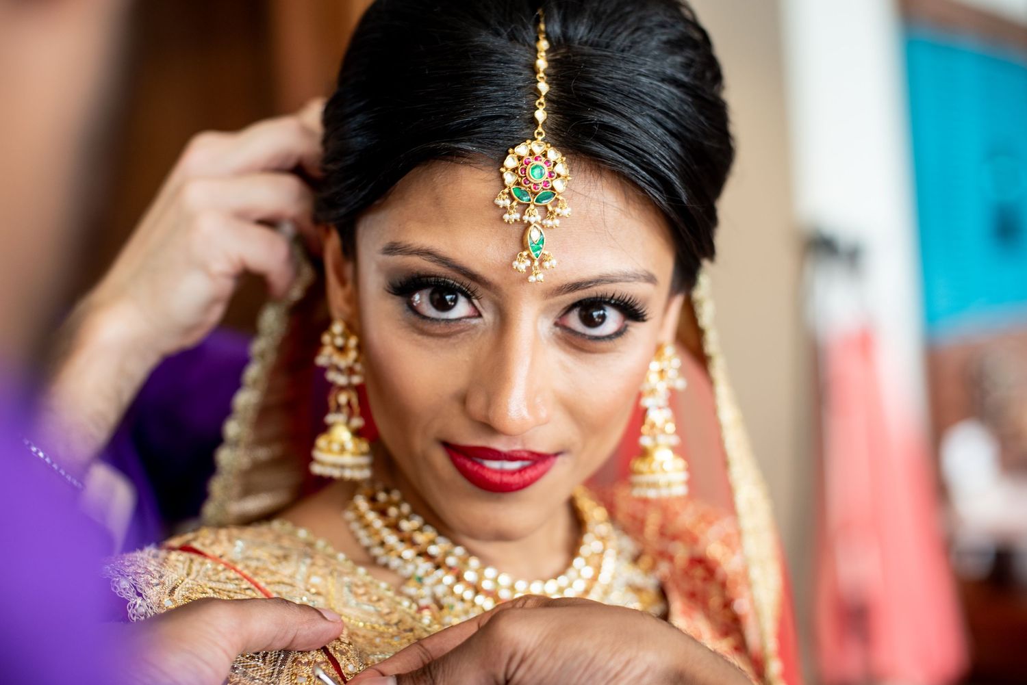 Indian bride in traditional gold jewelry and red lips getting ready, wearing ornate maang tikka and necklace.