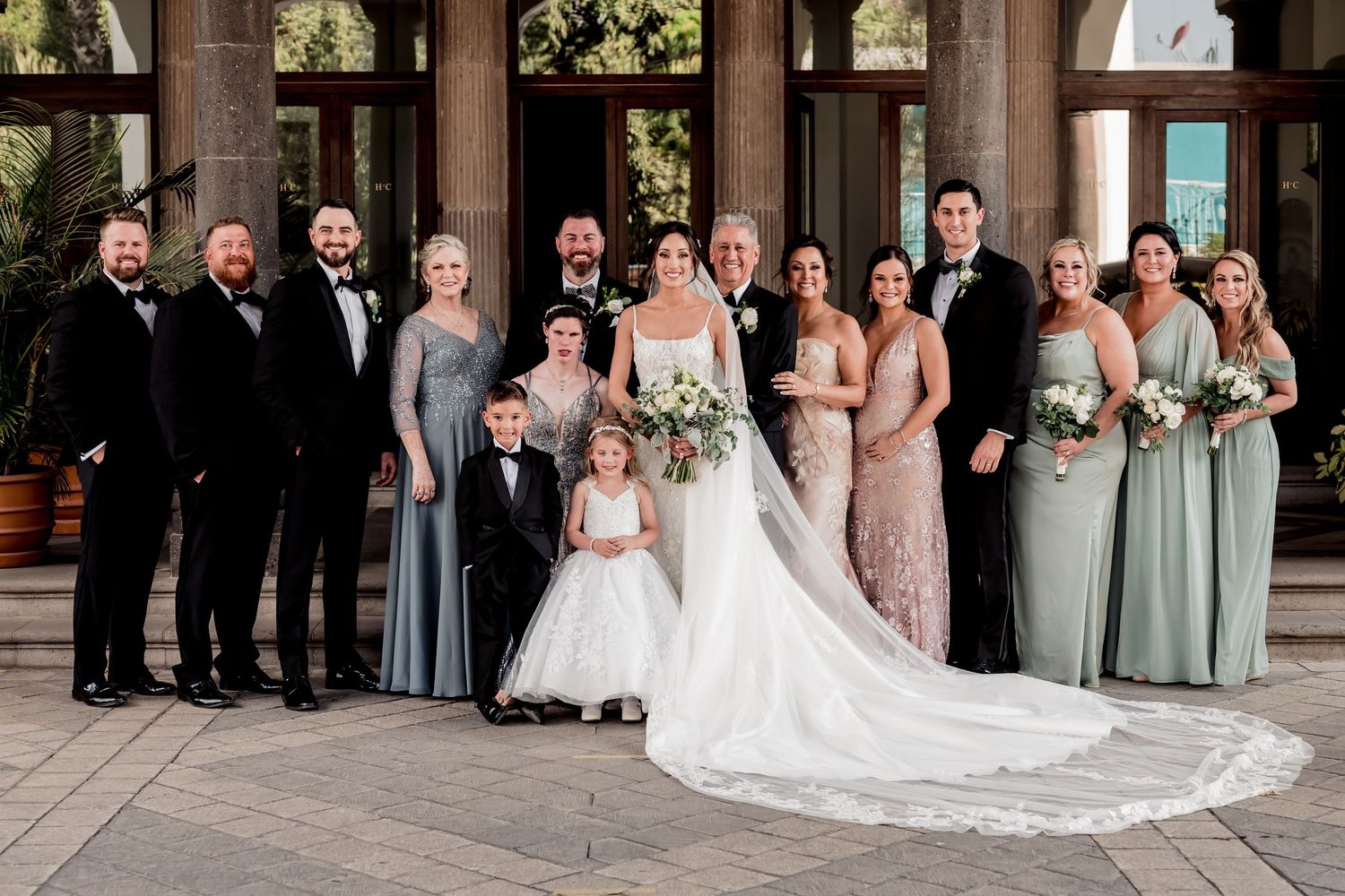 Wedding party poses in elegant attire outside a venue, bride in white gown with bouquet, bridesmaids in sage green dresses.