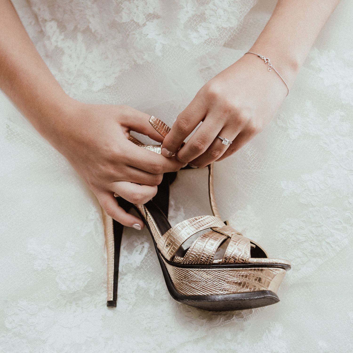 Close-up of hands fastening a metallic platform sandal with a white wedding dress in background.