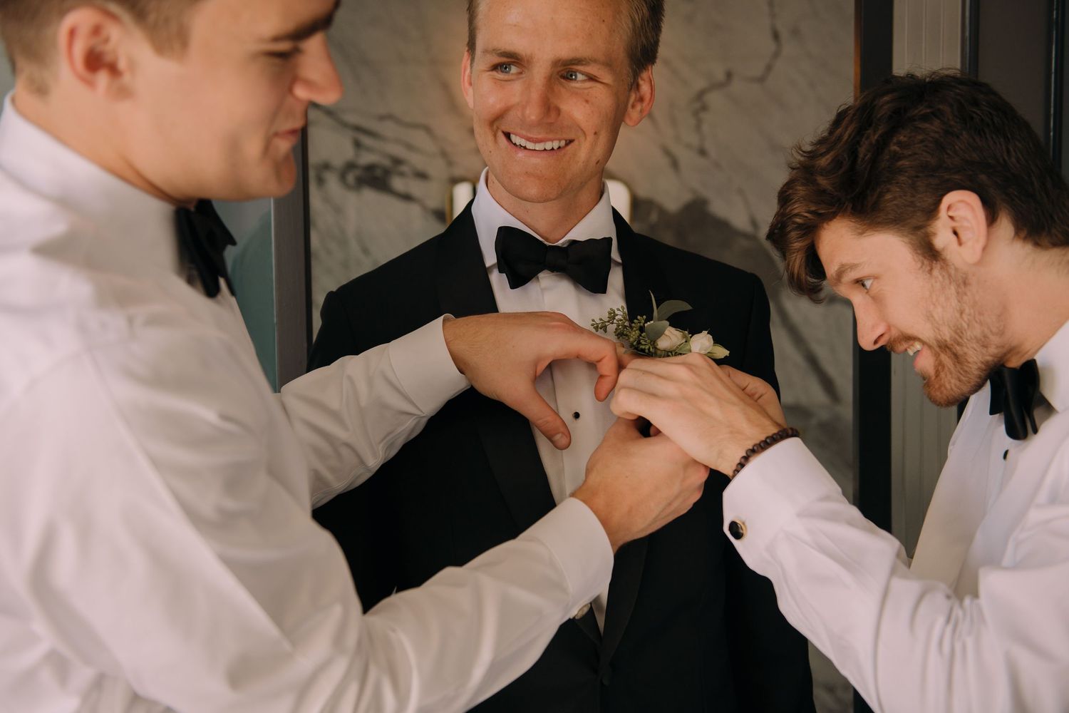 Groomsmen helping groom pin boutonniere on tuxedo lapel before wedding ceremony.