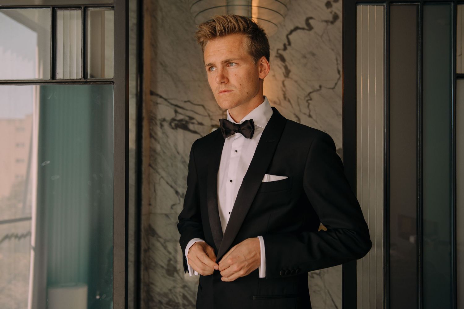 Elegant groom in black tuxedo with bow tie and white pocket square adjusting jacket in luxury venue.