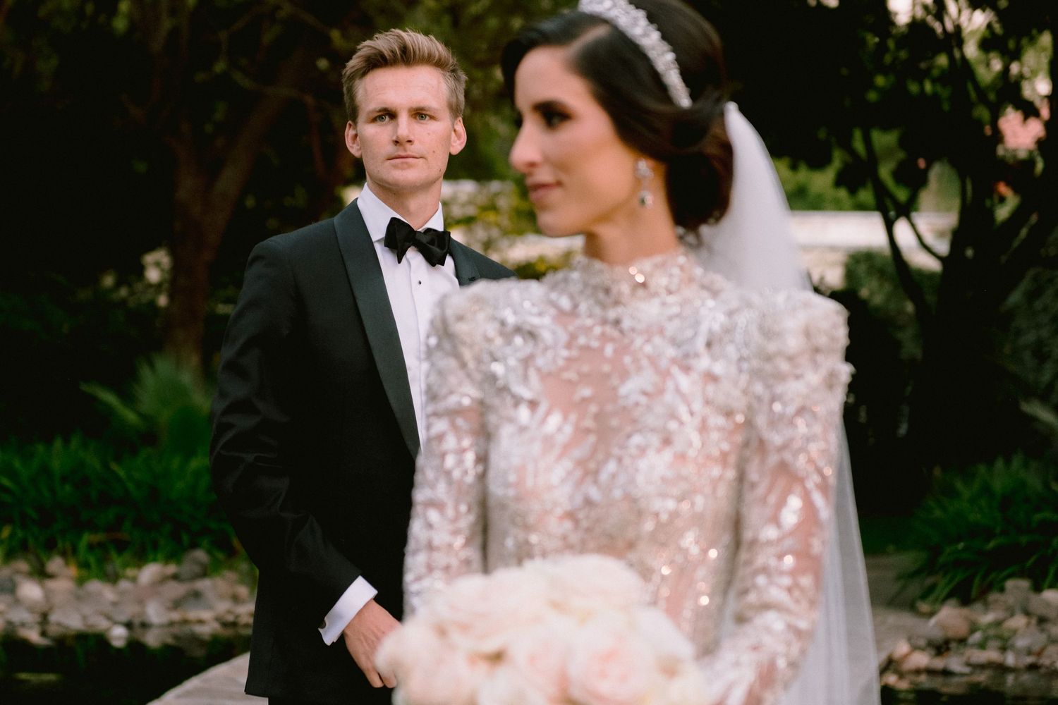 Bride in beaded long-sleeve gown with veil and groom in black tuxedo posing outdoors at elegant wedding.