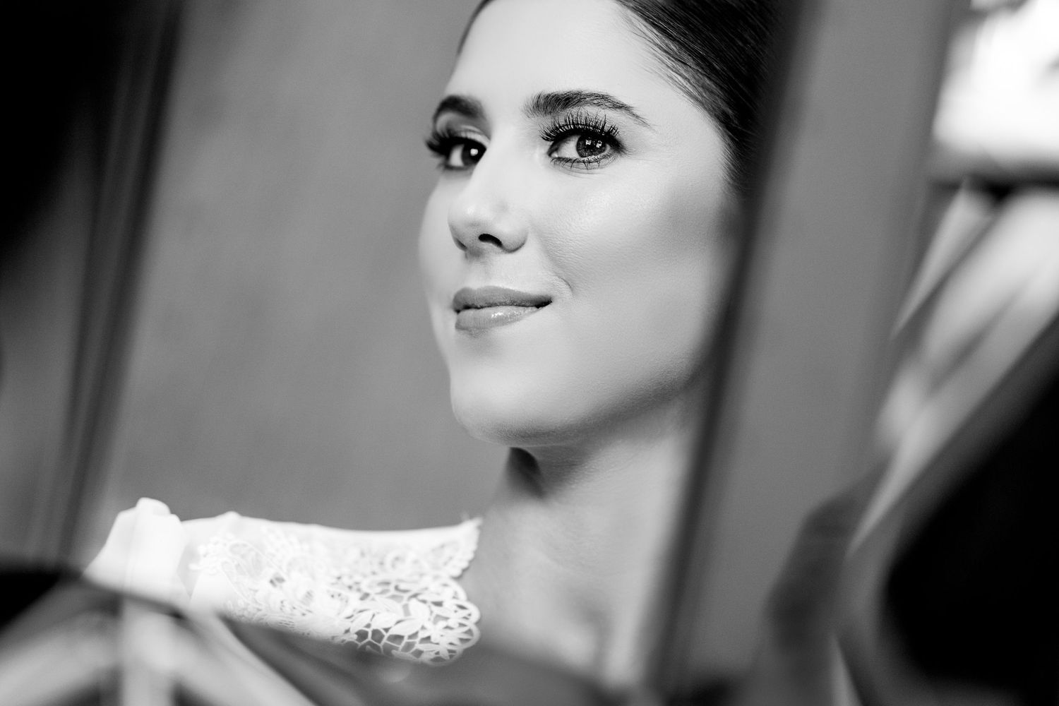 Black and white portrait of an elegant bride with dramatic eye makeup holding lace wedding dress in mirror reflection.