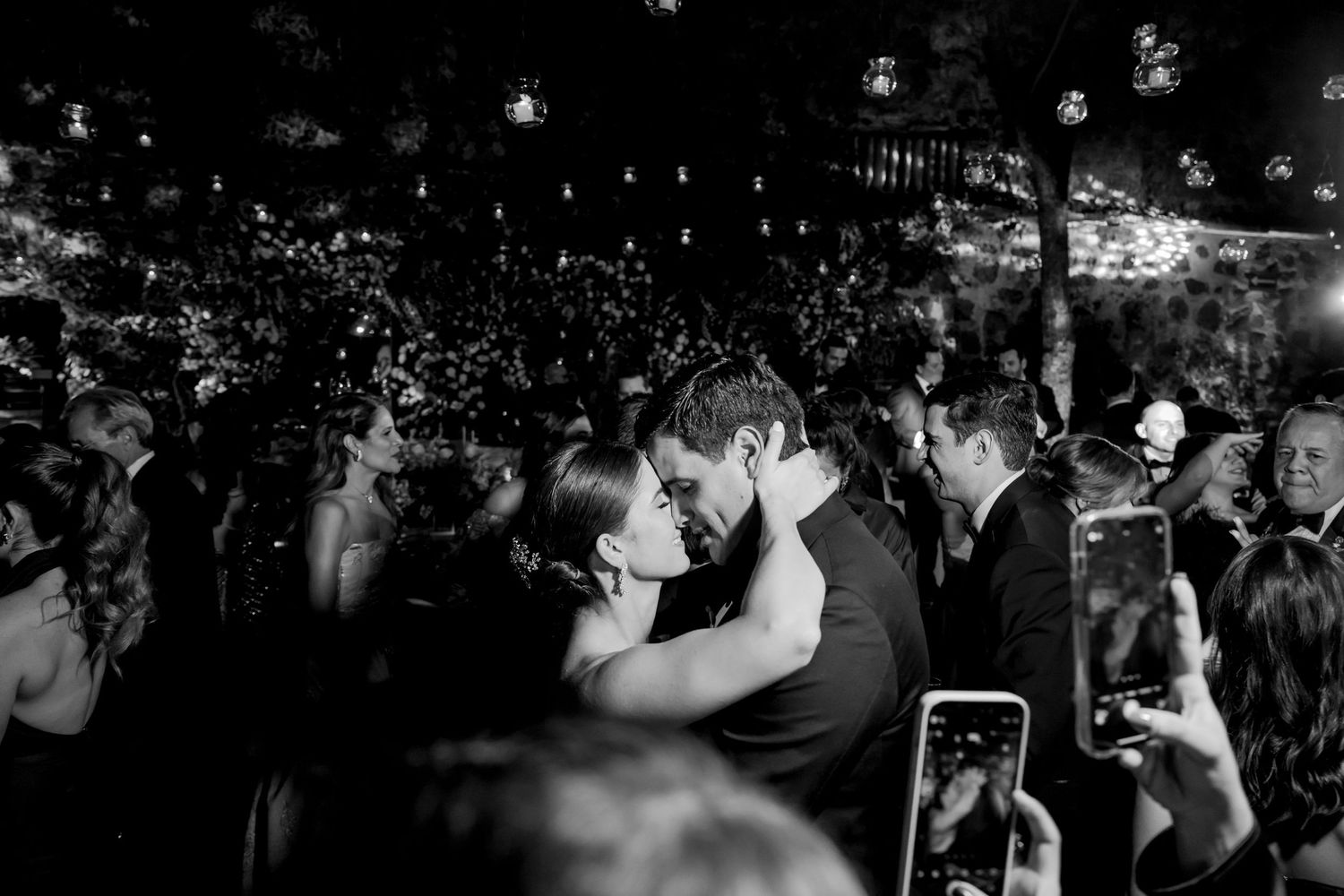 Black and white wedding photo of couple sharing romantic kiss surrounded by guests at outdoor evening reception.