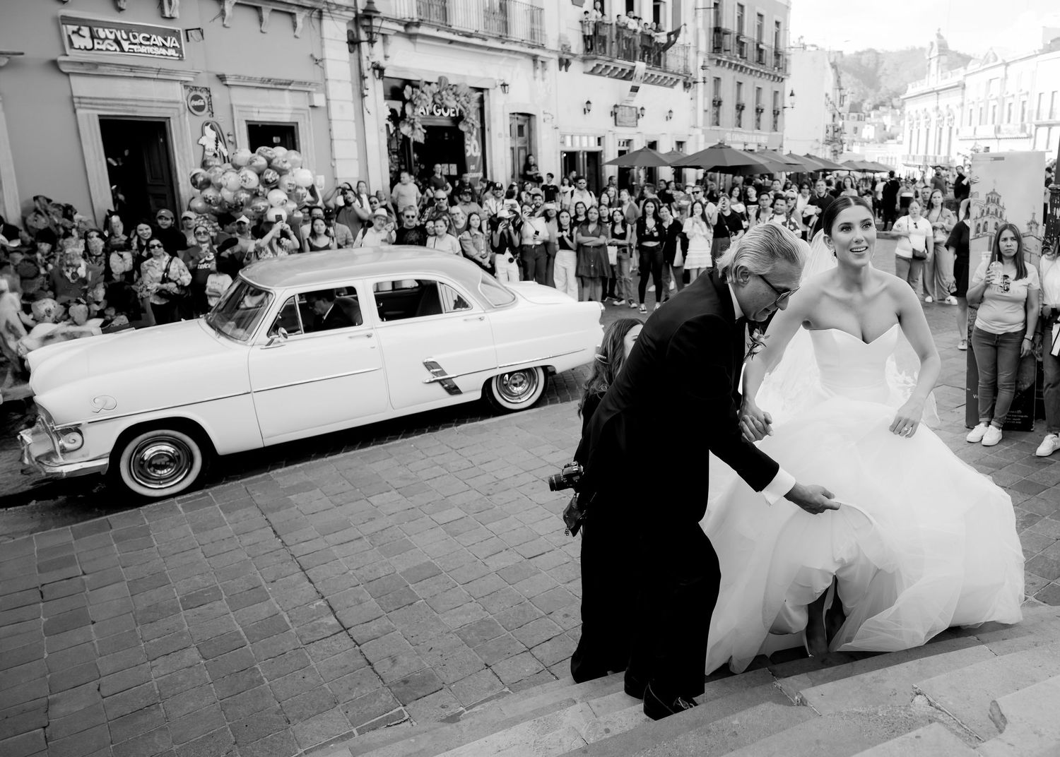 Black and white wedding photo of bride and groom arriving at church with vintage white car and crowd of guests.