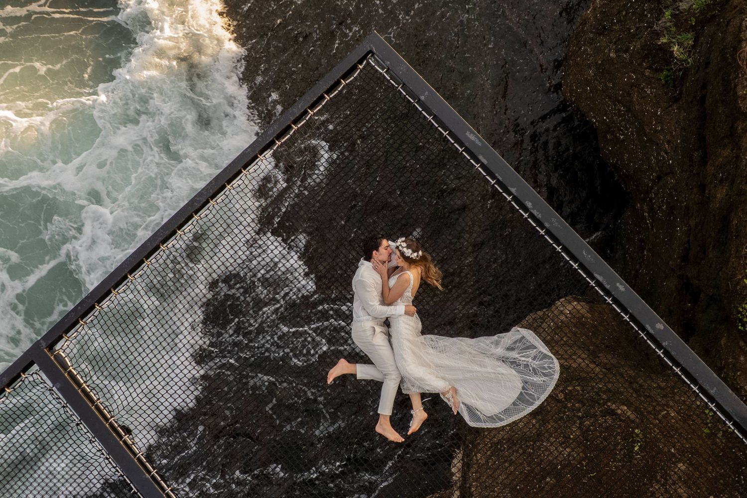 Aerial view of newlyweds sharing a romantic kiss on a coastal platform with ocean waves crashing nearby.