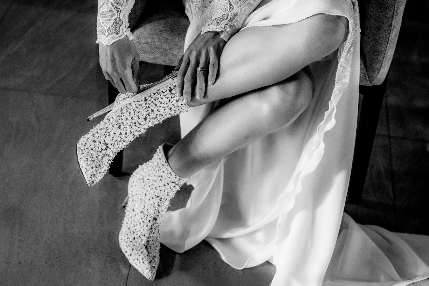 Black and white photo of a bride in a white lace gown putting on elegant lace heels while seated.