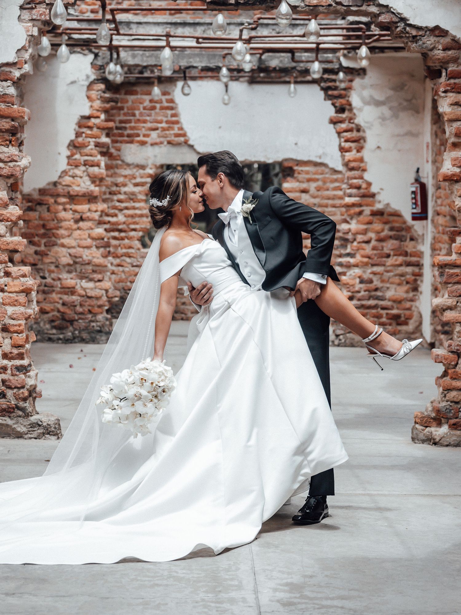 Romantic wedding couple dips and poses against rustic exposed brick wall in elegant formal attire.