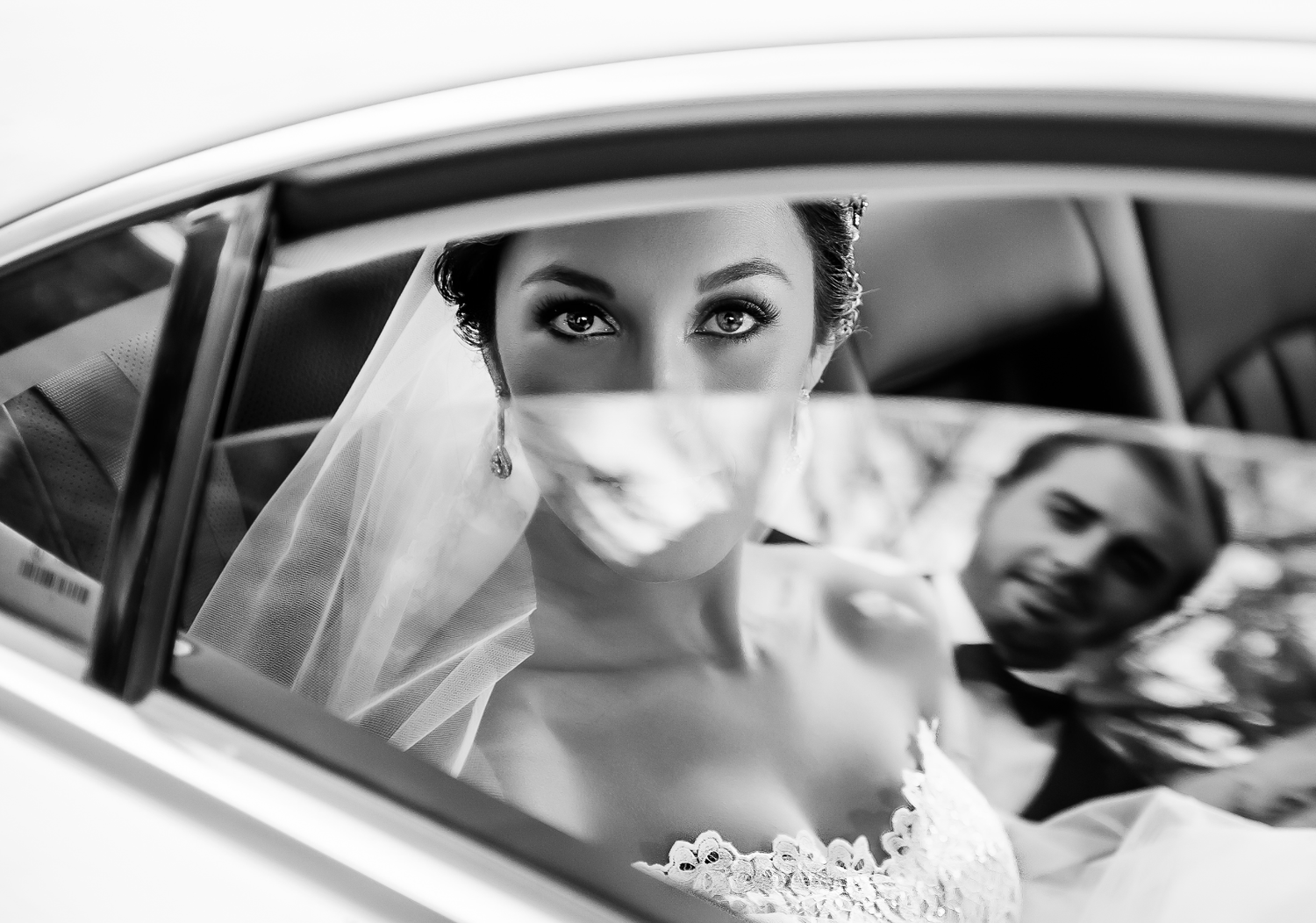 Black and white photo of a bride in a veil looking out a car window, with groom visible in reflection.