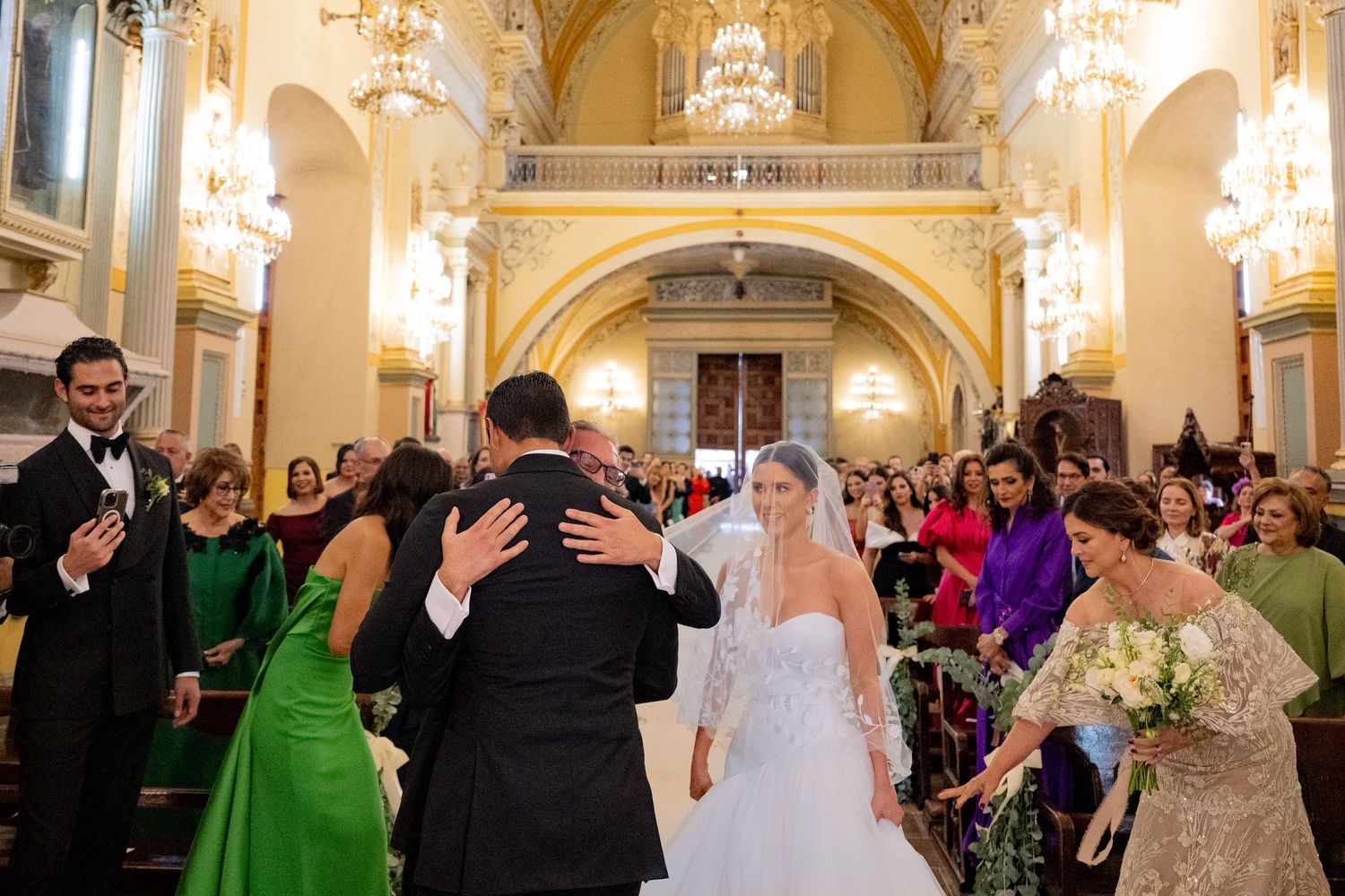 Bride and groom embrace at church altar during elegant wedding ceremony with guests in formal attire and ornate chandeliers.