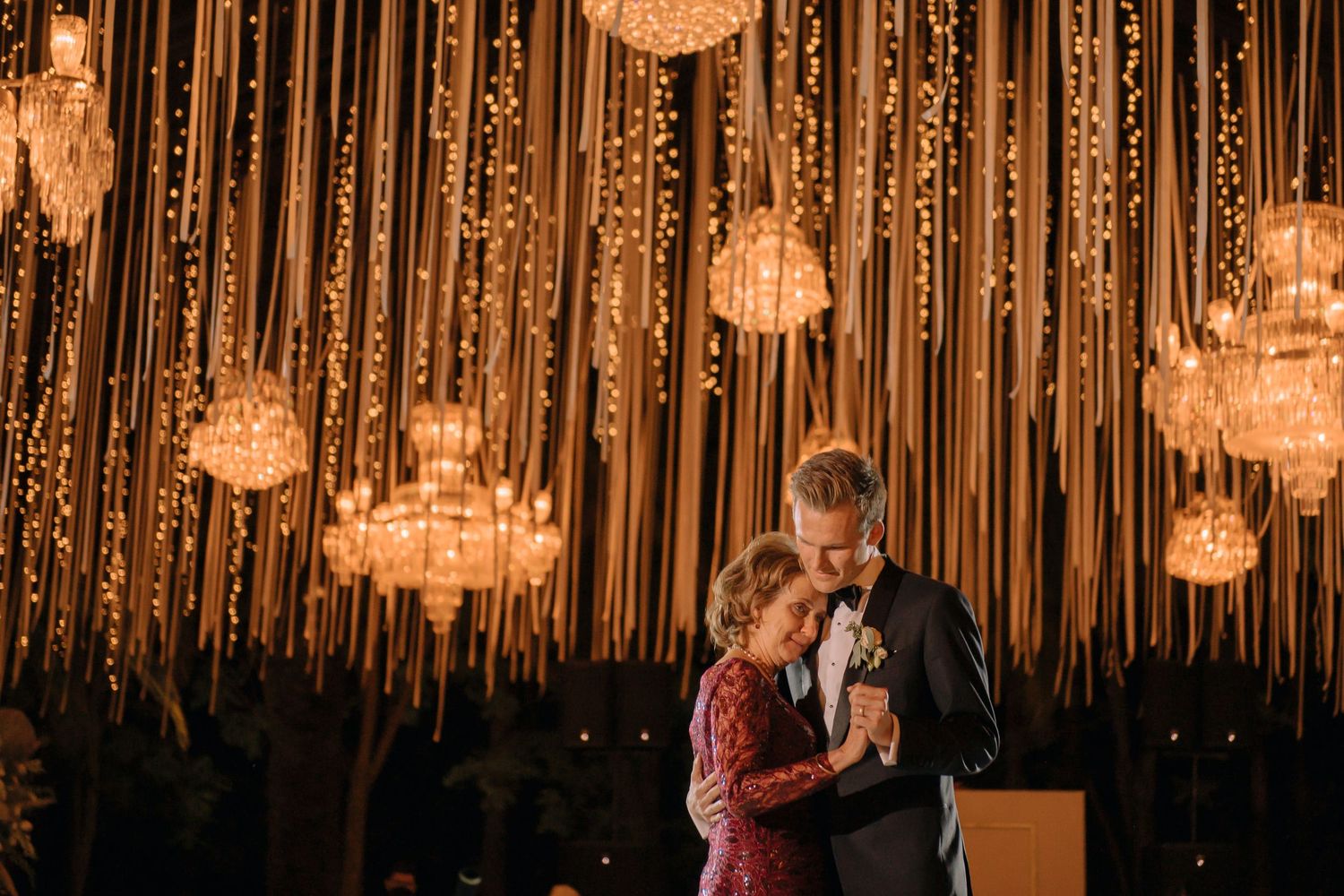 Couple shares first dance under crystal chandeliers and warm string lights at elegant wedding reception.