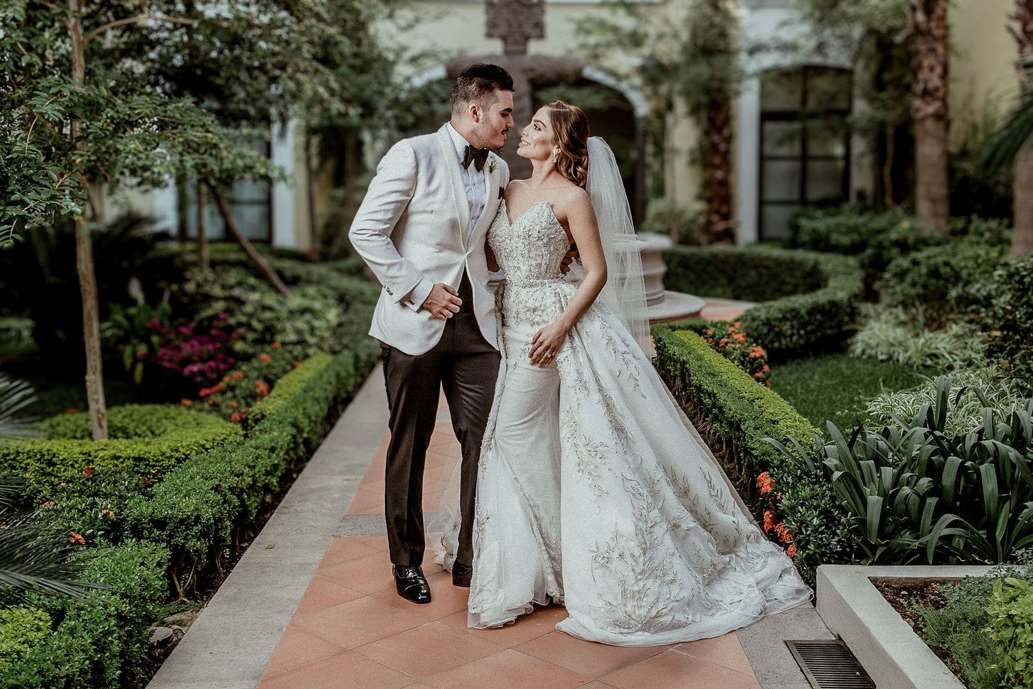 Elegant bride and groom share a romantic moment in a lush garden courtyard, bride wearing a stunning lace wedding gown.