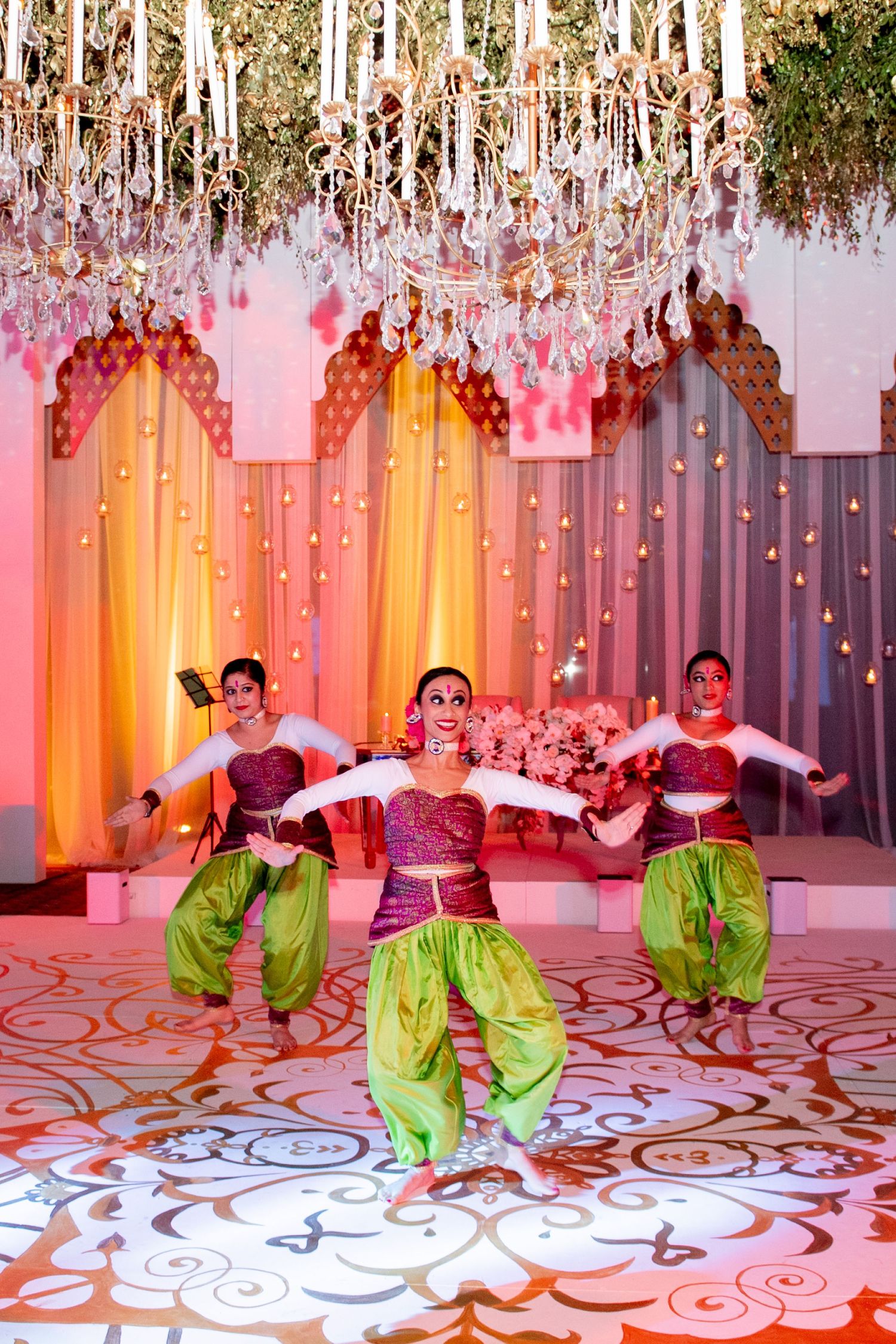 Vibrant Indian dancers in green and pink outfits perform on a decorated stage with crystal chandeliers at a festive event.