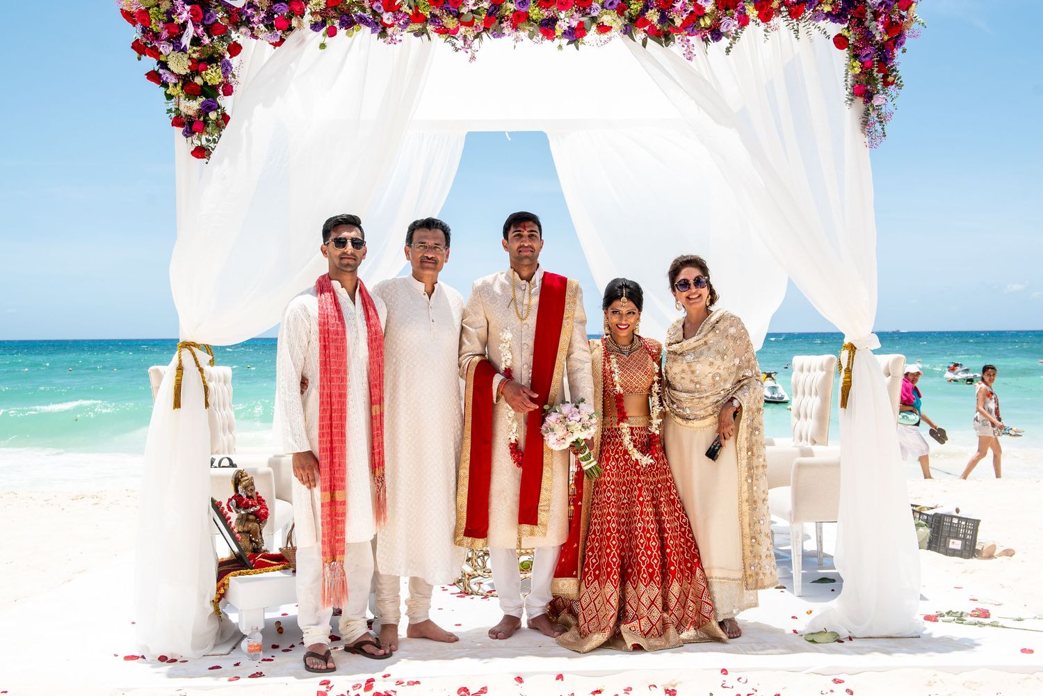 Indian wedding ceremony on a beach with five people under a floral arch, bride in red lehenga, blue sky backdrop.