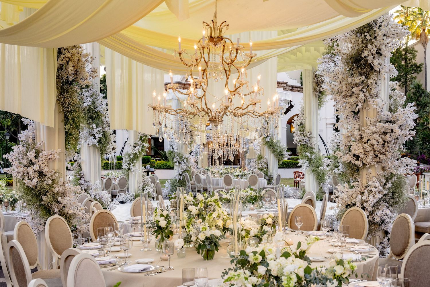 Elegant wedding reception with white floral arrangements, crystal chandelier, draped ceilings, and round tables with gold chairs.