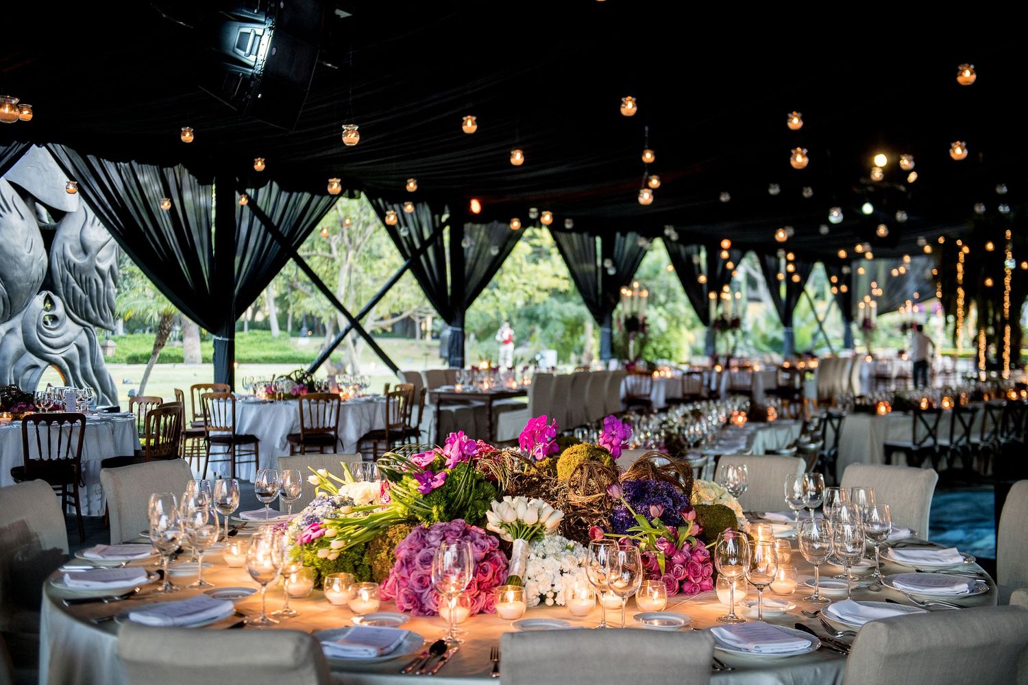 Elegant wedding reception tent with floral centerpieces, candlelight, string lights, and draped black fabric decor.